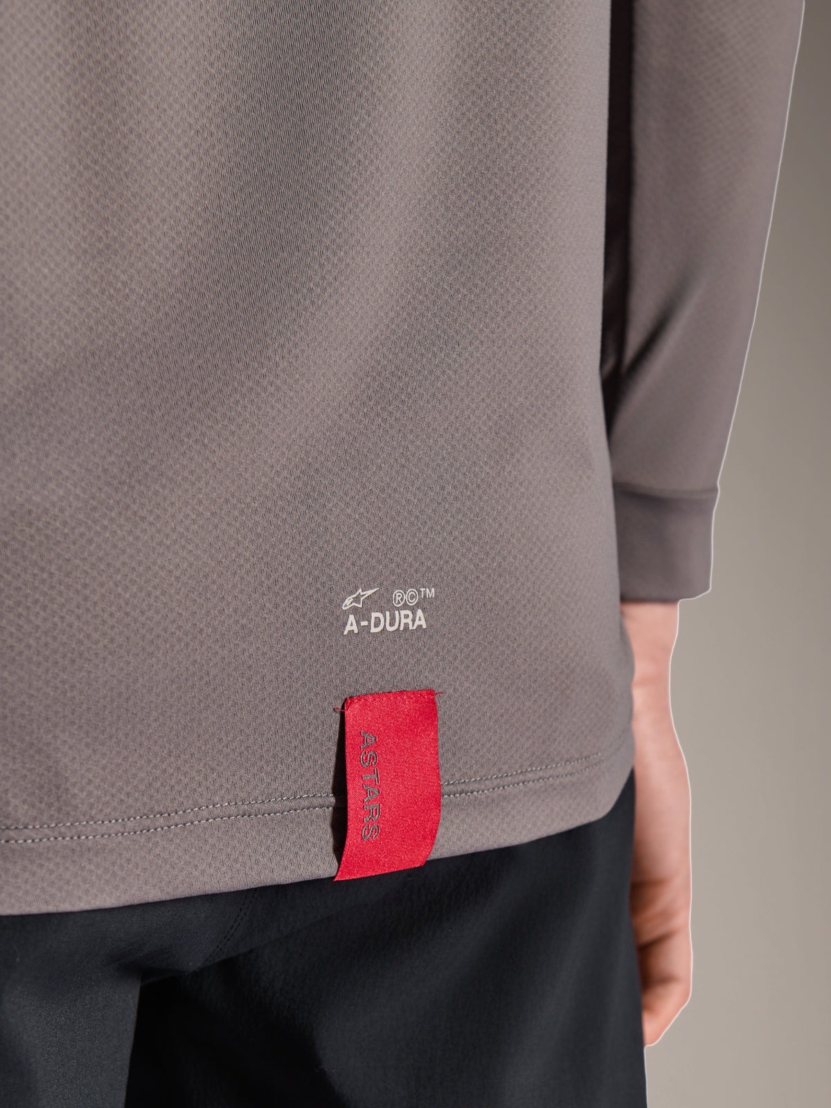 Alpinestars A-Dura Insert Astar Trikot, Langarm-MTB-Trikot, Dunkelgrau, Detailaufnahme des unteren Rückensaums mit weißem A-DURA-Branding und markantem roten ASTARS-Stoffetikett, strukturiertes Funktionsmaterial