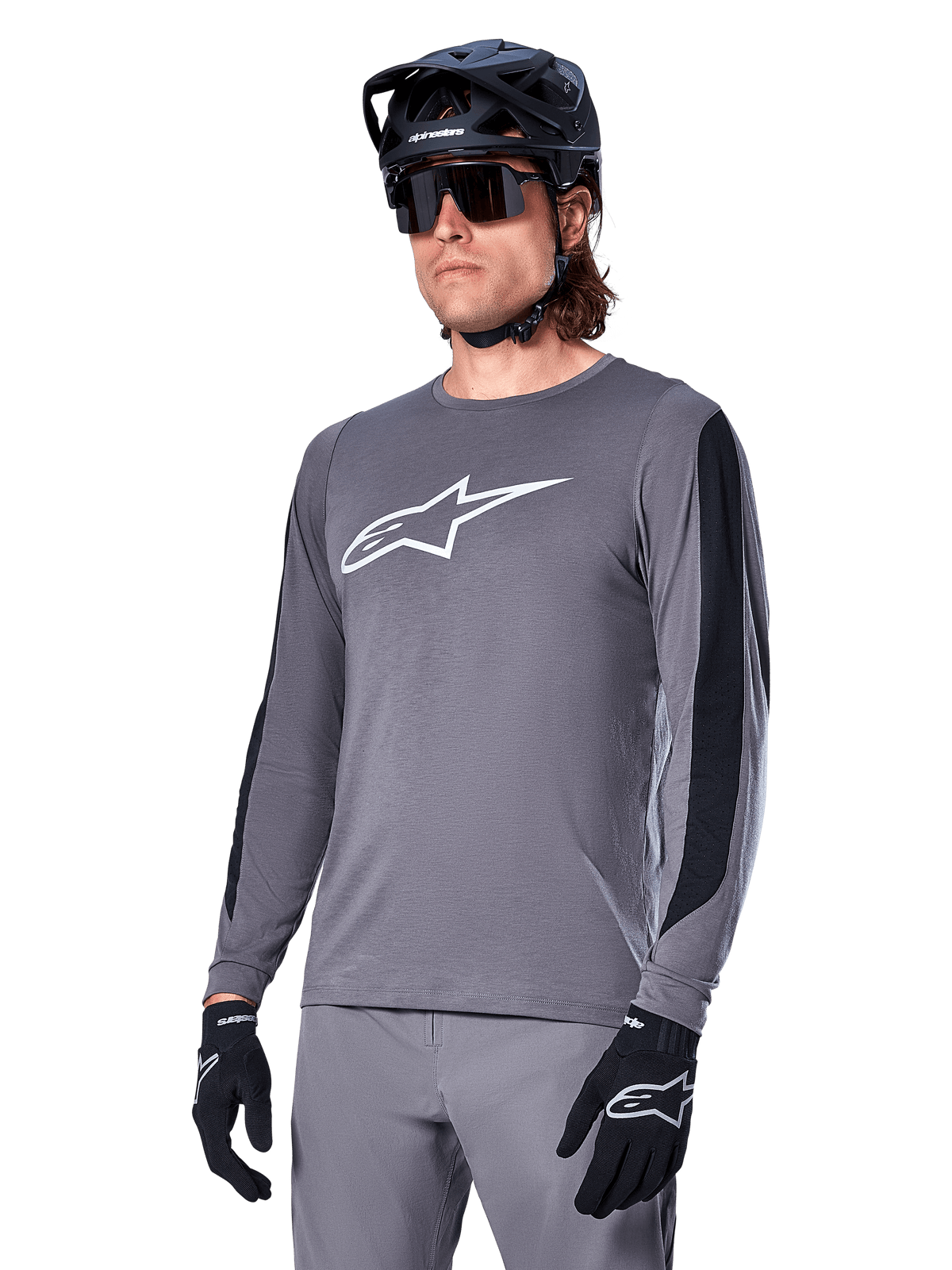 Alpinestars A-Dura Dri Astar Langarmtrikot, MTB-Mountainbike-Trikot, Dunkelgrau mit weißem Brustlogo und schwarzen Mesh-Seiteneinsätzen, getragen mit schwarzem Helm, Sonnenbrille und passender grauer Hose
