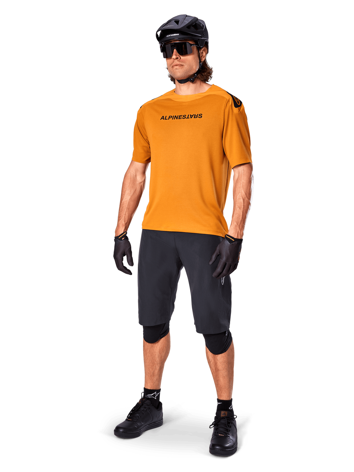 Männlicher Fahrer im Alpinestars A-Aria Polartec Switch Trikot in Dark Gold, kurzärmeliges MTB-Trikot, kombiniert mit schwarzen Radshorts, Knieschonern, schwarzem Helm und Handschuhen, professionelles Mountainbike-Outfit für Trail-Riding