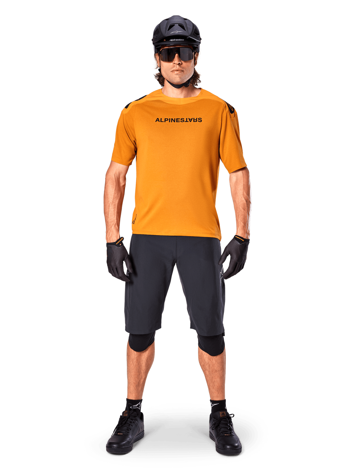 Männlicher Radfahrer im Alpinestars A-Aria Polartec Switch Kurzarmtrikot in Dark Gold, kombiniert mit schwarzen MTB-Shorts, Helm, Sonnenbrille und Handschuhen, professionelle Mountainbike-Trail-Ausrüstung