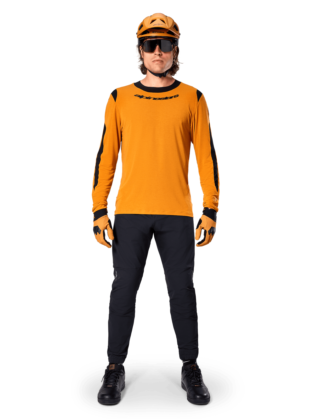 Mountainbiker im Alpinestars A-Dura Dri Wool Langarmtrikot, Dunkelgold mit schwarzen Akzenten an Kragen und Ärmeln, kombiniert mit einer schwarzen Hose, passendem goldenem Helm, Handschuhen und einer schwarzen Sonnenbrille, professionelle Mountainbike-Ausrüstung