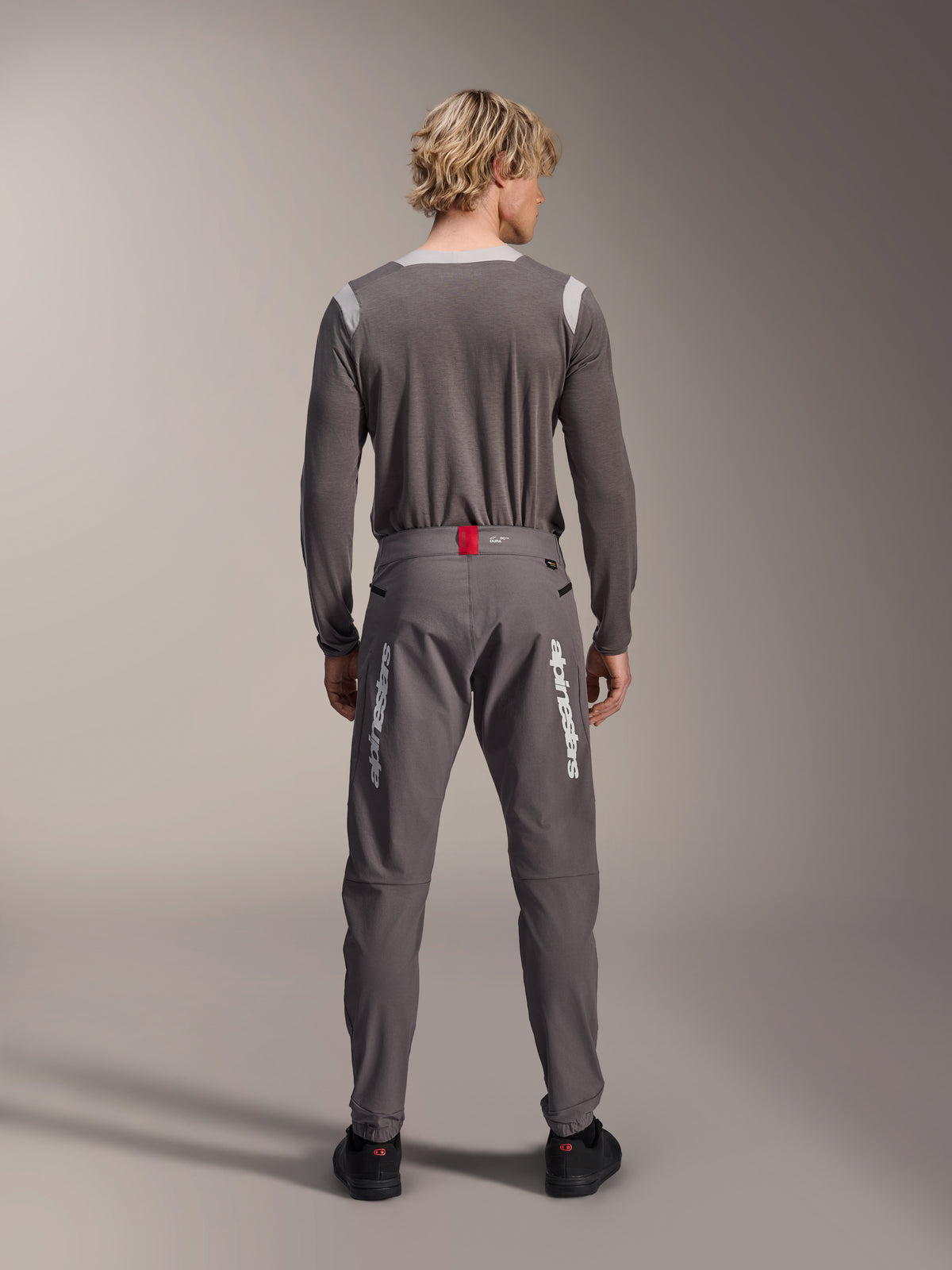 Model in der Rückansicht trägt Alpinestars A-Dura Elite Hose in Dunkelgrau und ein passendes graues Langarm-Trikot, weiße Alpinestars-Logos auf den Beinen, roter Akzent am Bund, schwarze Schuhe, professionelle MTB-Ausrüstung.