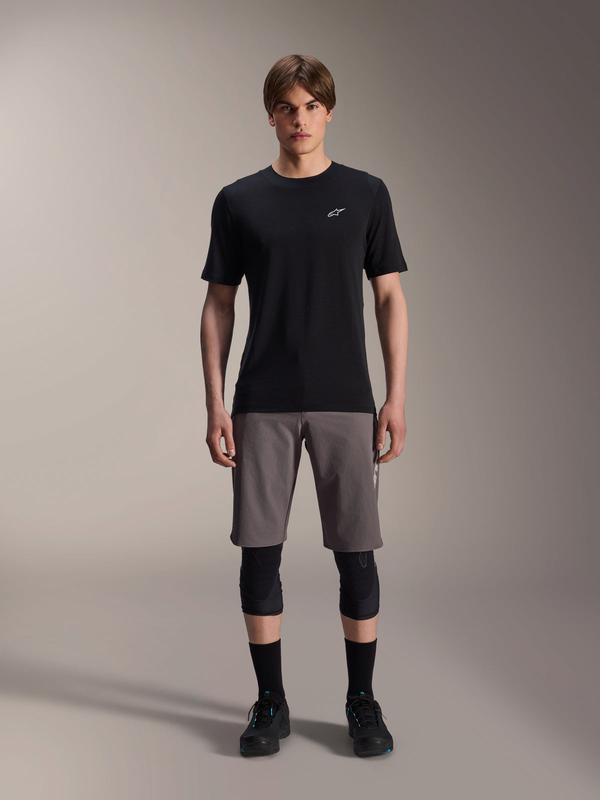 Männliches Model trägt Alpinestars A-Dura Elite Shorts in Dunkelgrau und das A-Dura Dri-Wool Kurzarm-Trikot in Schwarz, kombiniert mit schwarzen Knieschonern und Radschuhen; technische Mountainbike-Bekleidung für Offroad-Performance.