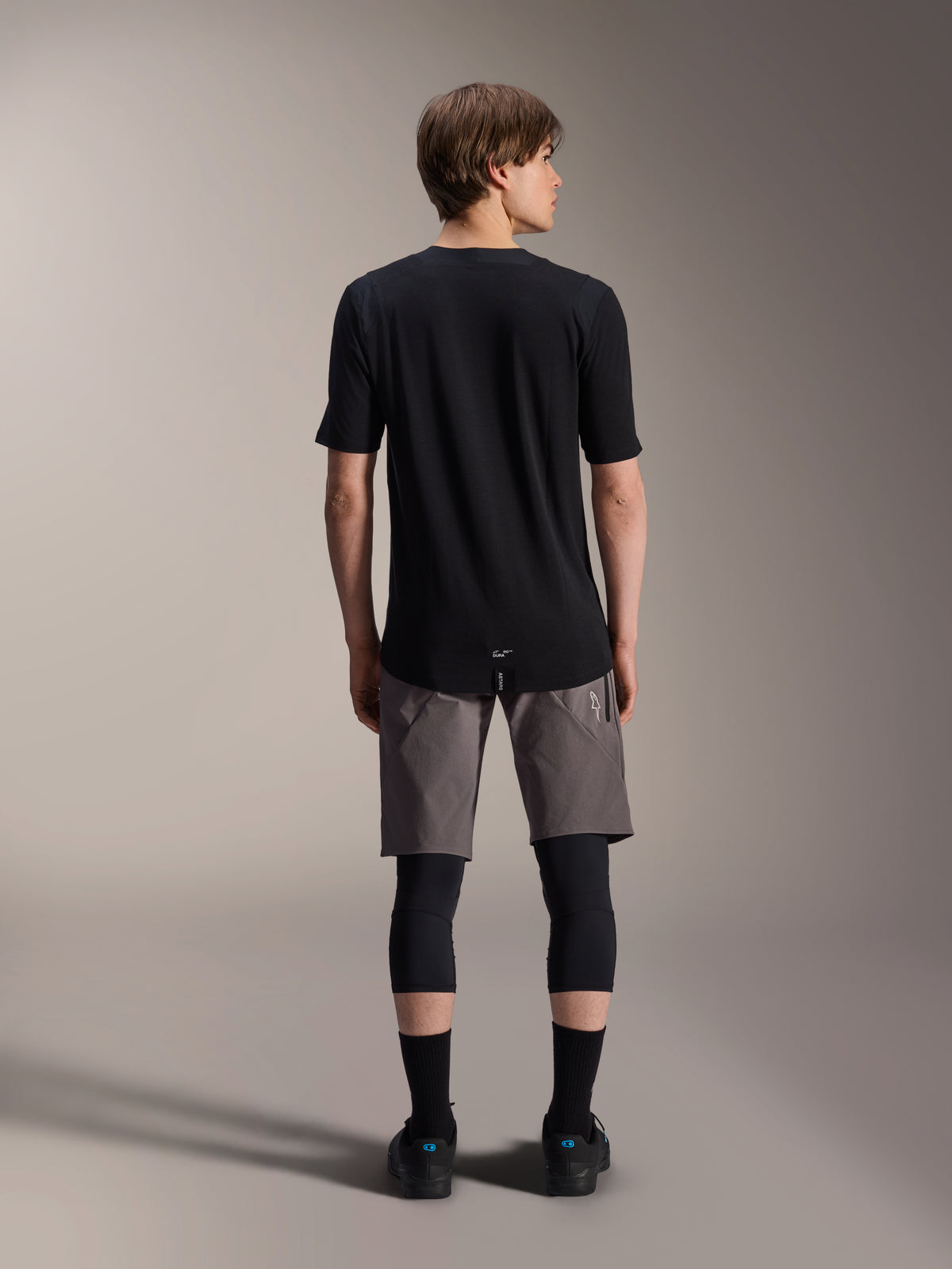 Alpinestars A-Dura Dri Wool Kurzarmtrikot, Schwarz, Rückansicht getragen von einem Modell mit grauen MTB-Shorts und schwarzen Knieschützern, mit längerem Rückensaum und athletischer Passform für den Offroad-Radsport