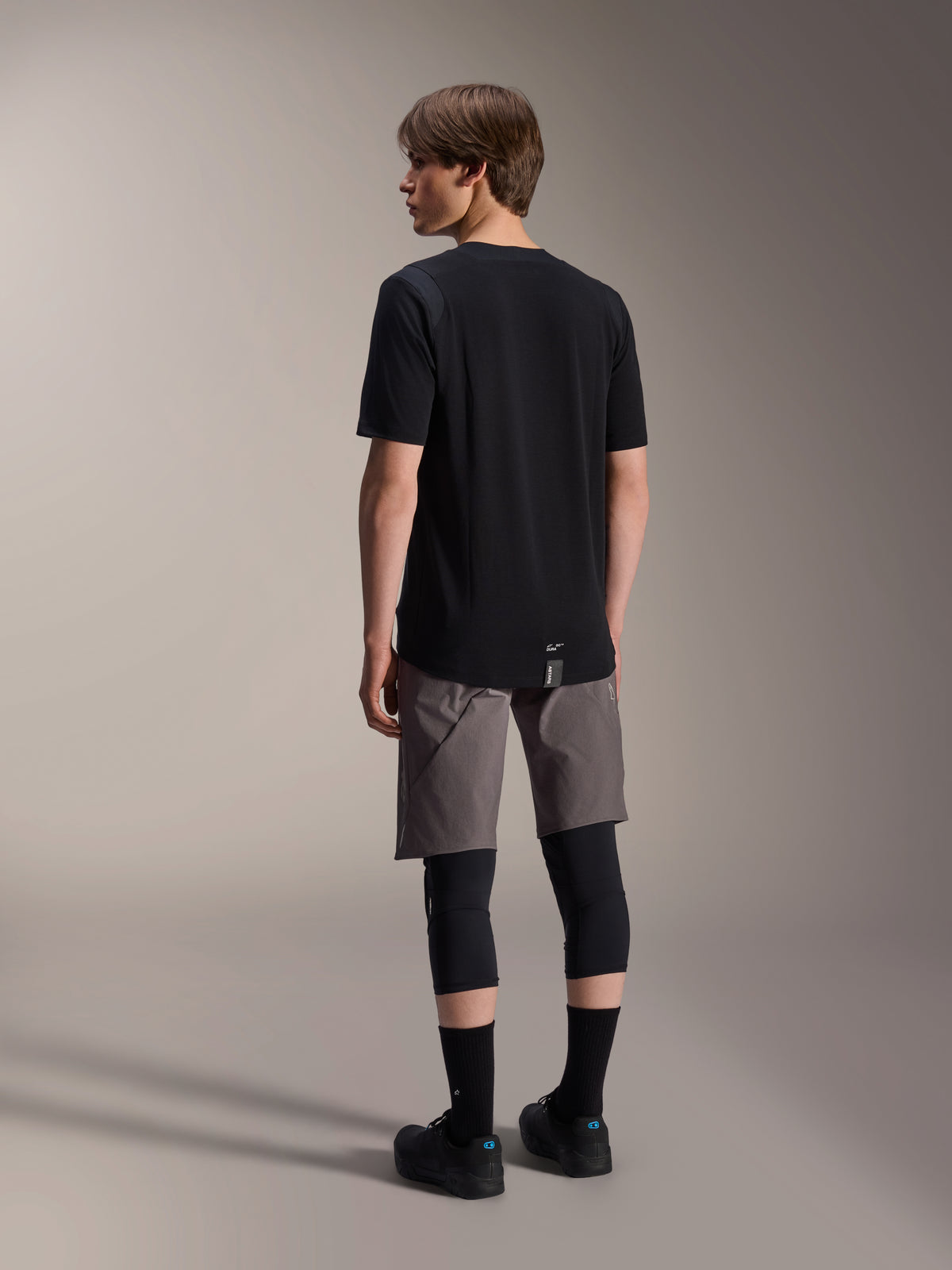 Alpinestars A-Dura Dri Wool Kurzarmtrikot und A-Dura Elite Shorts, schwarz- und dunkelgraue Mountainbike-Bekleidung, getragen mit schwarzen Knieschützern und Socken, Rückansicht der Radsportausrüstung für Offroad-Performance