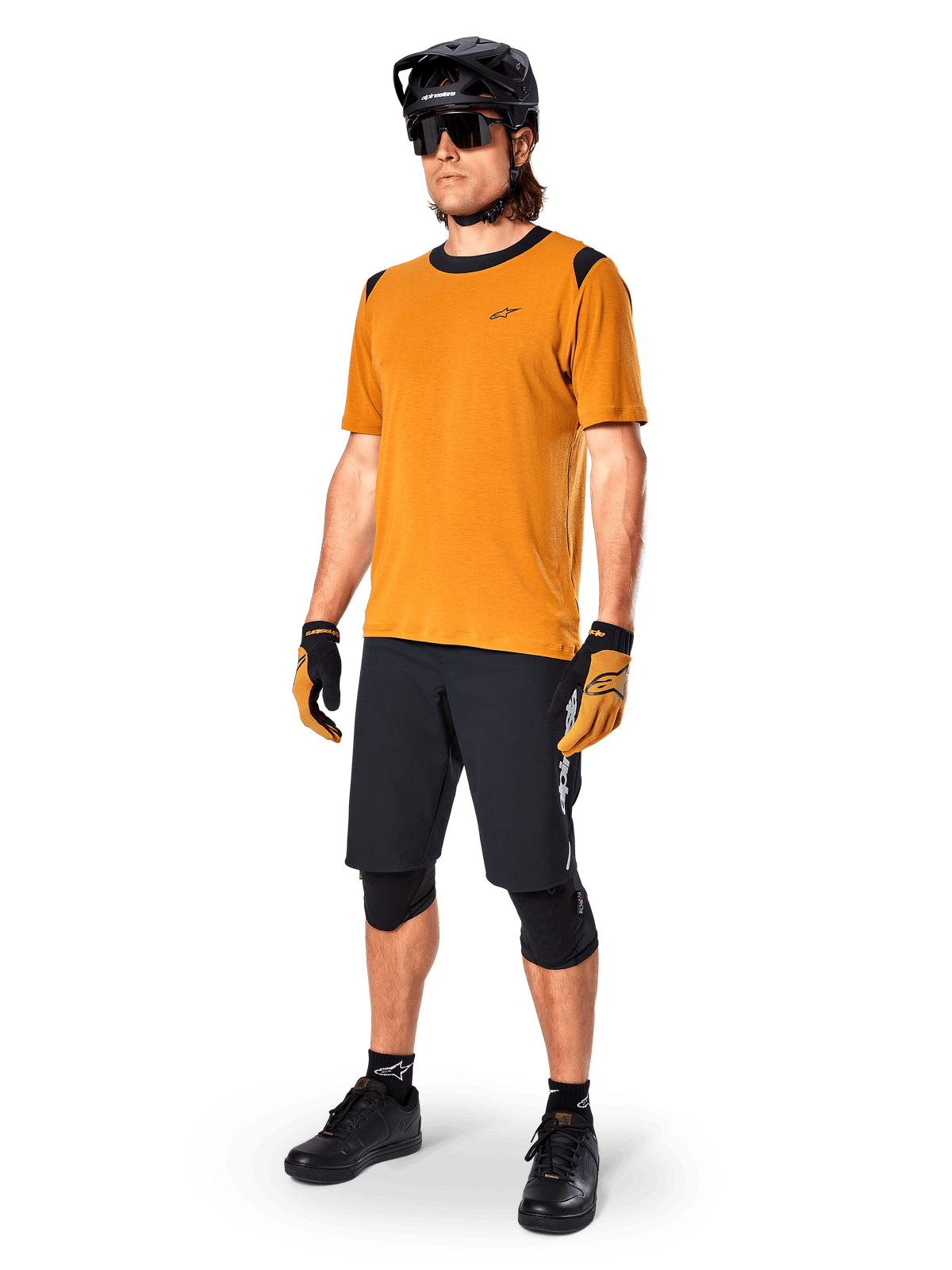 Male model wearing Alpinestars A-Dura Dri Wool Jersey - Short Sleeve in Dark Gold, mountain bike jersey with black shoulder and collar accents, paired with black MTB shorts, black helmet, and sunglasses, technical off-road cycling gear