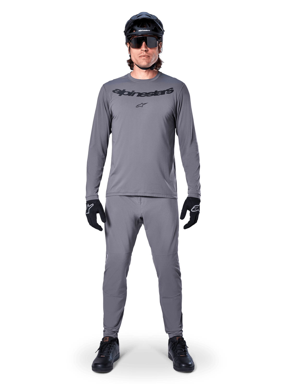 Male cyclist wearing Alpinestars A-Dura Rocker Long Sleeve Jersey in Dark Gray, matching gray pants, black MTB helmet, sunglasses and gloves, professional mountain bike apparel for off-road trails