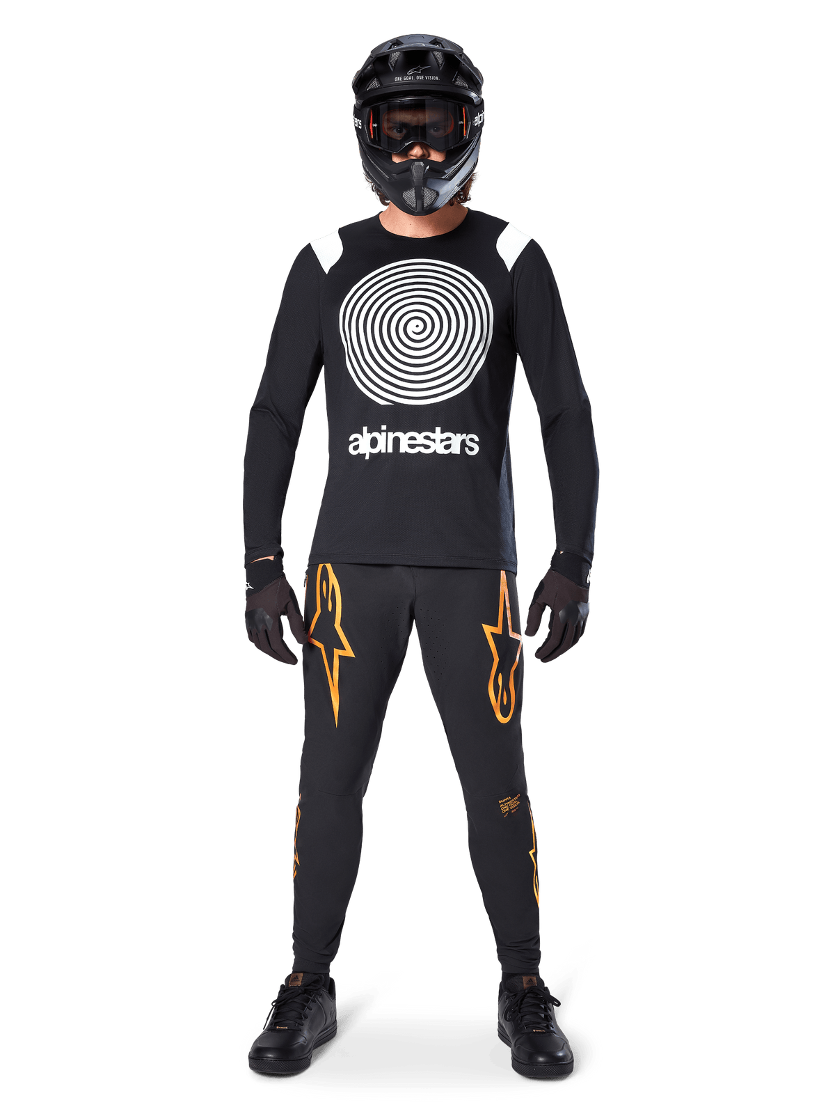 Männliches Model in kompletter Mountainbike-Ausrüstung, Alpinestars A-Supra Oscar Langarm-Trikot in Schwarz mit weißer Spiralgrafik auf der Brust, schwarze Hose mit orangefarbenen Stern-Logos, schwarzer Fullface-Helm und Goggle, schwarze Handschuhe und Schuhe, koordiniertes Outfit für MTB-Gravity-Sport