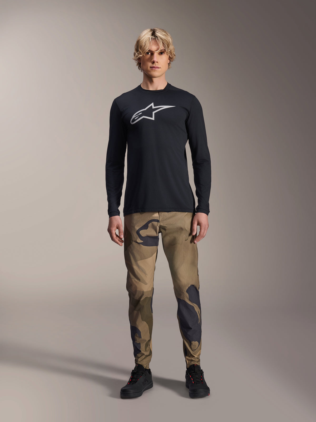 Alpinestars A-Dura Astar Langarmtrikot, Schwarz, mit einem großen weißen Astars-Logo auf der Brust, getragen mit einer Camouflage-Hose und schwarzen Schuhen, lässige Mountainbike-Performance-Bekleidung