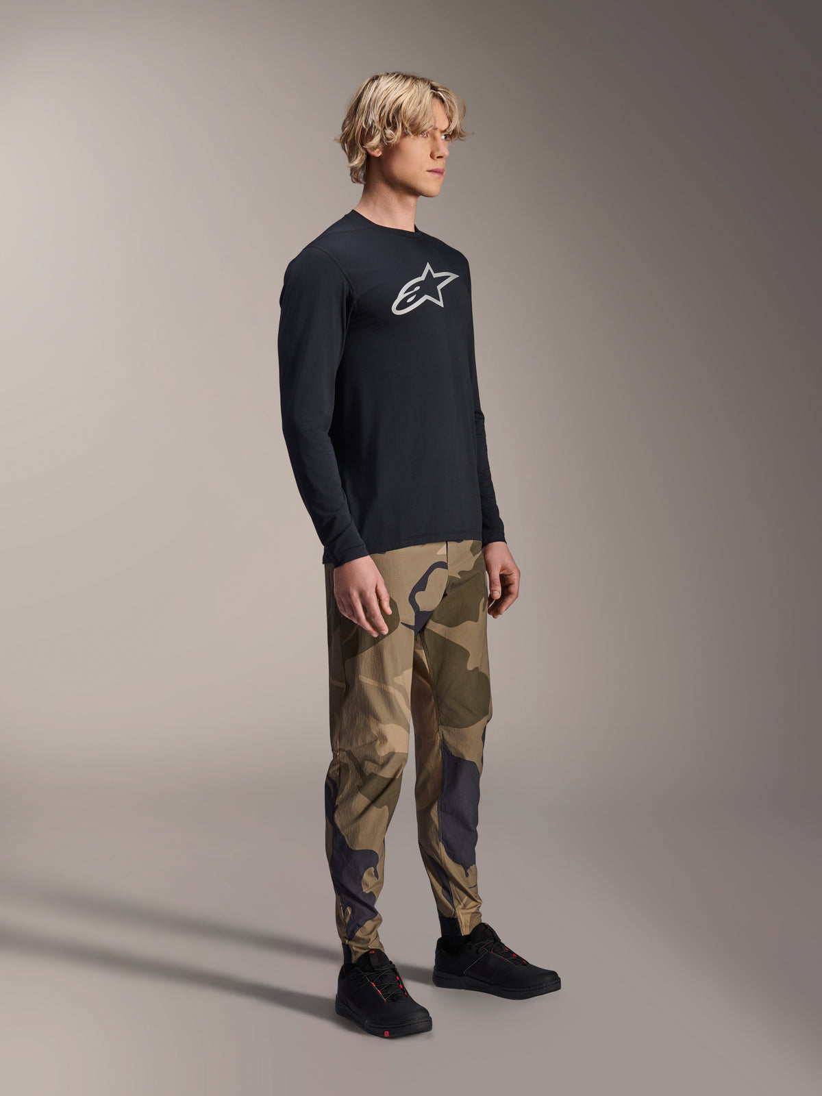 Alpinestars A-Dura Astar Langarm-Trikot in Schwarz, getragen von einem Modell mit Camouflage-Hose und schwarzen Sneakern, weißes Astars-Brustlogo, lässige Performance-MTB-Bekleidung für Trailriding