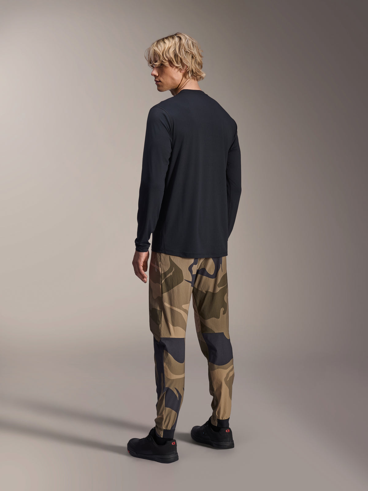 Alpinestars A-Dura Astar Langarmtrikot, Schwarz, Rückansicht an einem Model, kombiniert mit einer Camouflage-Hose, lässige MTB-Performance-Ausrüstung mit verlängertem Rückensaum und regulärer Passform