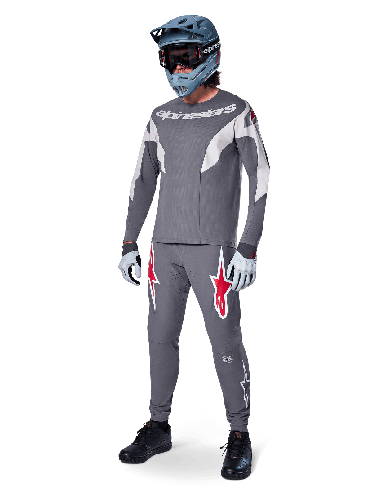 MTB-Fahrer im Alpinestars A-Supra Race Trikot, Langarm, Dunkelgrau mit hellgrauen Akzenten, passende dunkelgraue Hose mit rotem Stern-Logo, grauer Fullface-Helm mit Goggles, hellgraue Handschuhe, abgestimmtes Outfit für Mountainbike-Rennen
