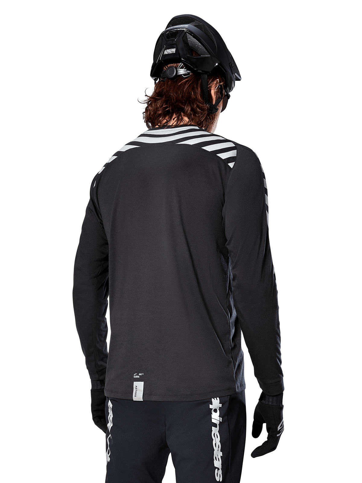Alpinestars A-Dura Dri Oscar Langarmtrikot, Black Reflective, Rückansicht mit reflektierenden Diagonalstreifen auf den Schultern und einer verlängerten Rückenpartie, getragen mit schwarzem MTB-Helm und Handschuhen, technische Ausrüstung für Mountainbiking