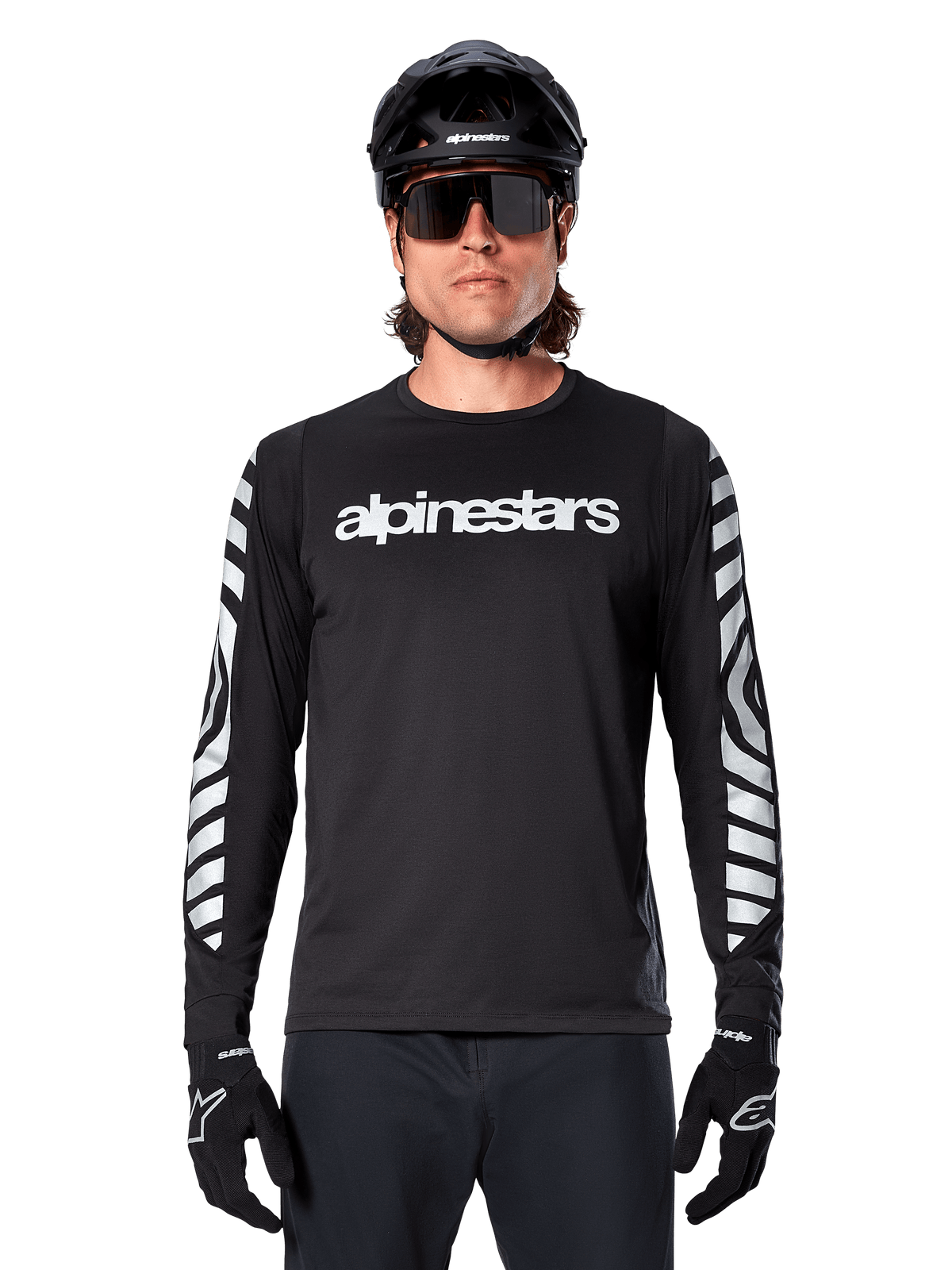 Männlicher Fahrer im Alpinestars A-Dura Dri Oscar Langarmtrikot, Schwarz Reflektierend, mit weißem Alpinestars-Schriftzug auf der Brust und silbernen reflektierenden Grafiken auf den Ärmeln, kombiniert mit schwarzem MTB-Helm, Sonnenbrille und schwarzen Handschuhen, Performance-Radsportbekleidung