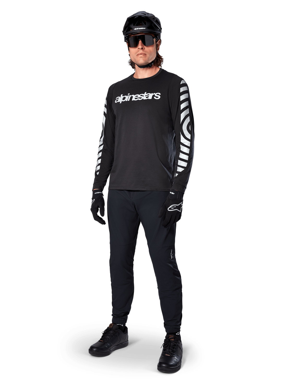 Alpinestars A-Dura Dri Oscar Langarmtrikot, Black Reflective, mit auffälligem weißem Alpinestars-Schriftzug auf der Brust und reflektierenden Grafikmustern auf den Ärmeln, kombiniert mit schwarzer Mountainbike-Hose, Helm und Handschuhen, technische MTB-Bekleidung