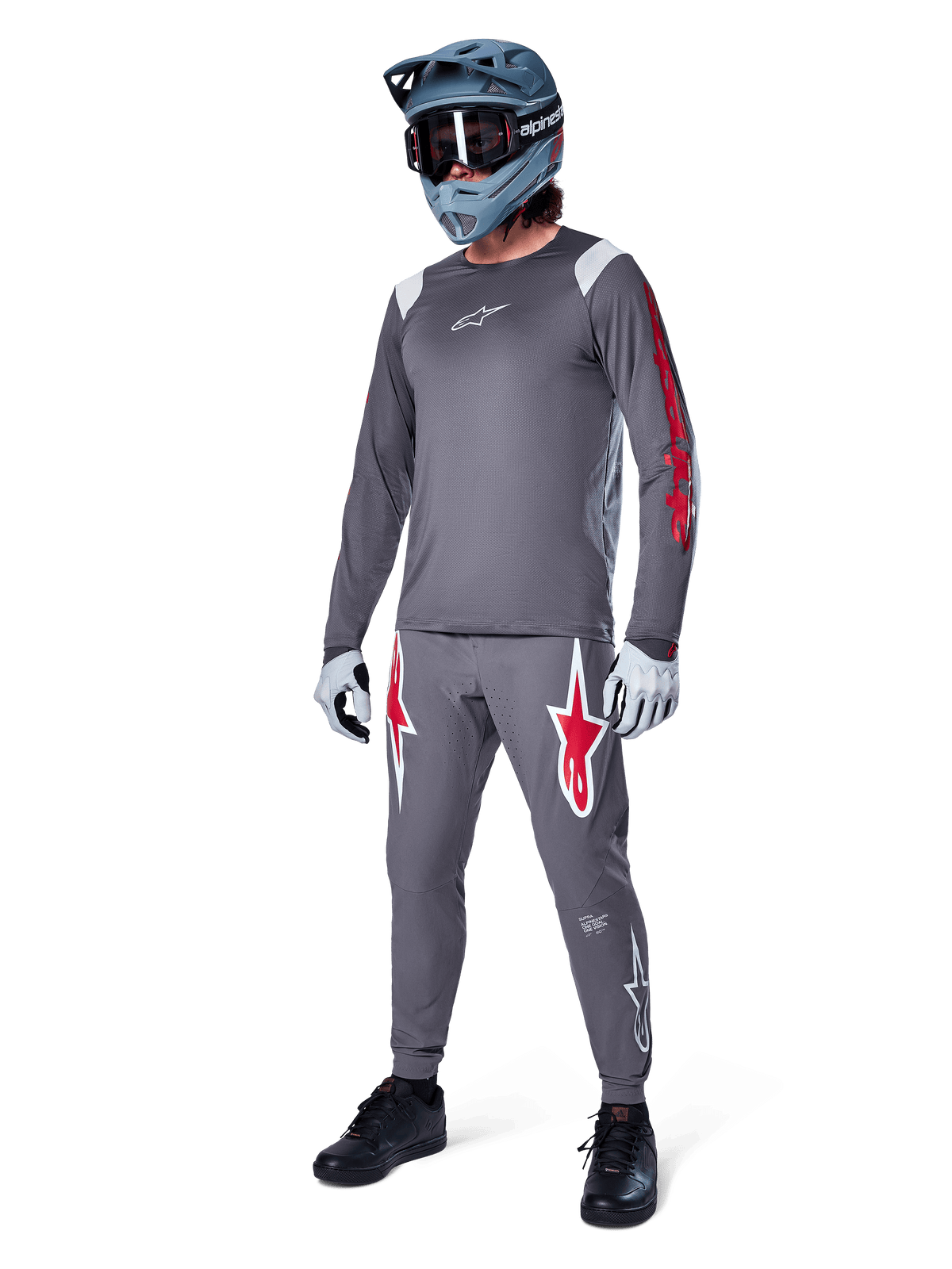 Alpinestars A-Supra Astar Langarmtrikot in Dunkelgrau, mit rotem Branding auf den Ärmeln und weißem Logo auf der Brust, getragen von einem Fahrer mit passender grauer Hose, einem blaugrauen Full-Face-Helm und Performance-Handschuhen, koordinierte Mountainbike-Ausrüstung für Gravity-Riding.