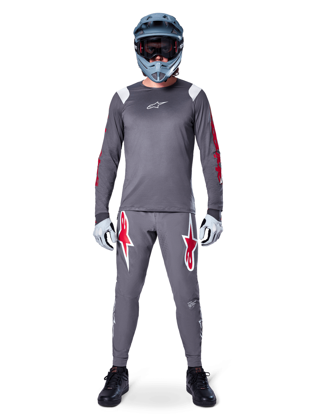 MTB-Fahrer im Alpinestars A-Supra Astar Langarmtrikot, Dunkelgrau mit roten Akzenten und weißem Brust-Logo, dazu passende graue Hose mit großen roten Sterngrafiken, blauer Fullface-Helm mit Goggle, hellgraue Handschuhe und schwarze Schuhe, professionelle Mountainbike-Ausrüstung