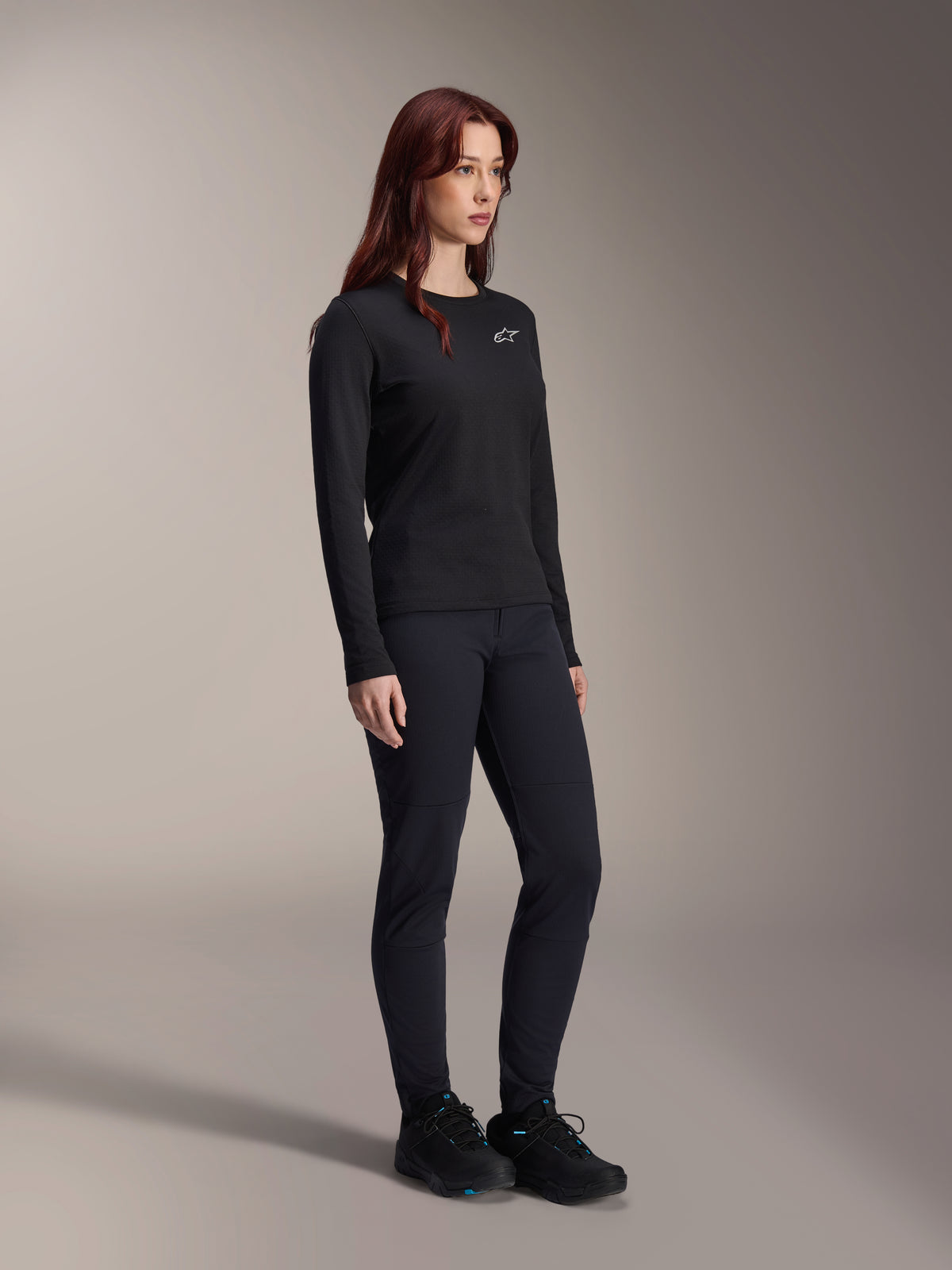 Woman wearing the Alpinestars Stella A-Dura Thermal Jersey, long-sleeve black MTB jersey with white chest logo, styled with matching black pants and athletic shoes, studio portrait for performance cycling apparel