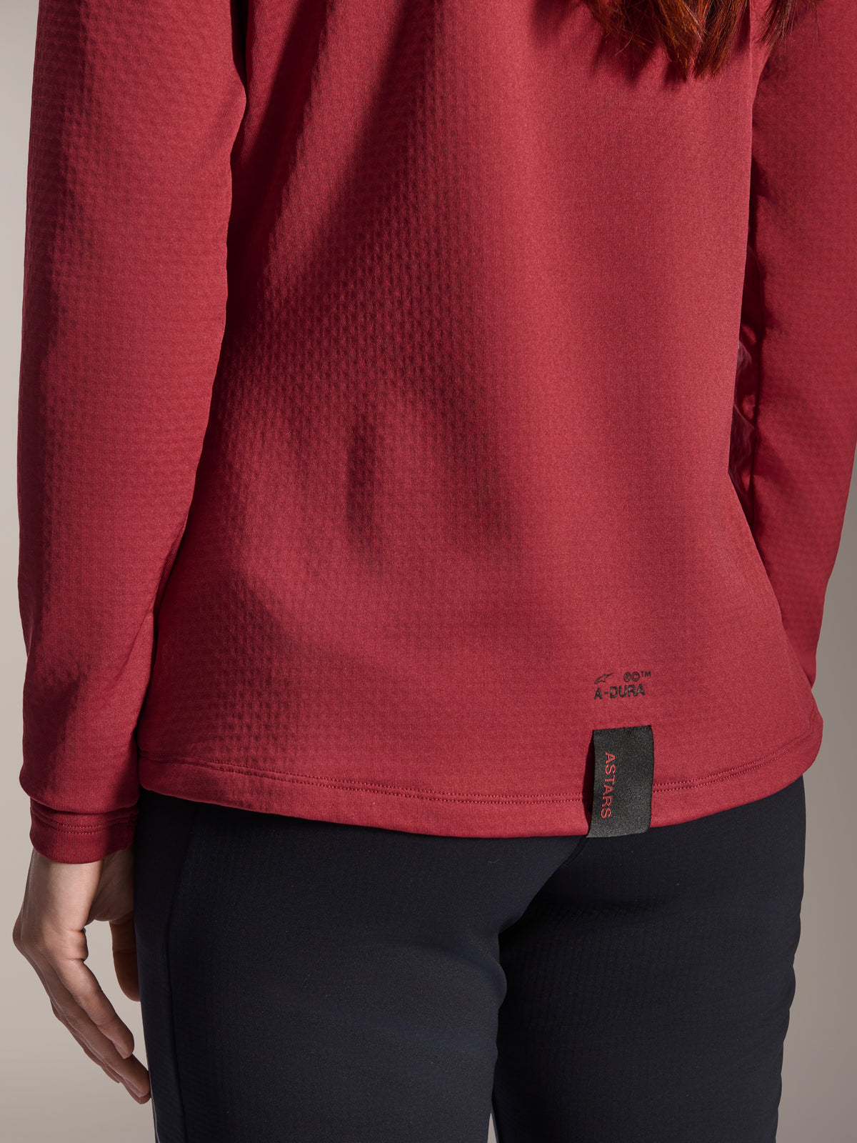 Alpinestars Woman Stella A-Dura Thermal Jersey, Burgundy, close-up view of the back hem featuring A-Dura branding and ASTARS woven tag, textured insulated fleece fabric, women's mountain bike apparel