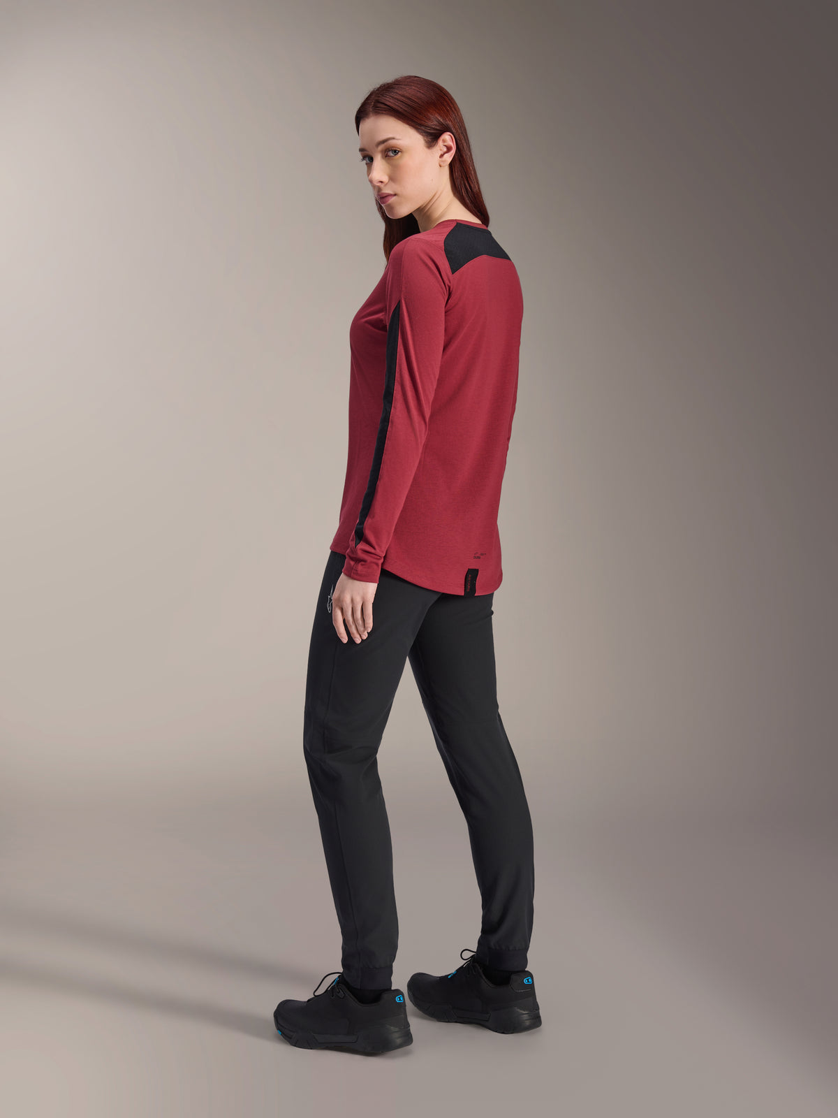Alpinestars Stella A-Dura Dri Astar Langarmtrikot für Damen, Burgunderrot und Schwarz, kombiniert mit schwarzer MTB-Hose und schwarzen Schuhen, Rückseiten-Profilansicht, recyceltes drirelease-Gewebe mit Mesh-Einsätzen, technische Mountainbike-Bekleidung