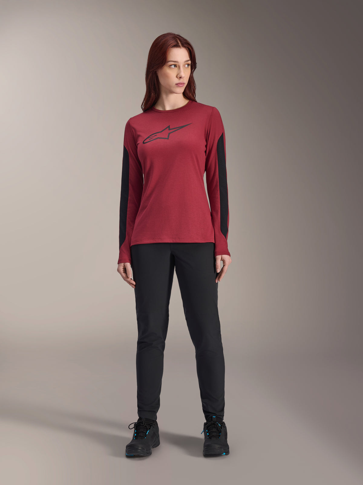 Alpinestars Stella A-Dura Dri Astar Langarmtrikot für Damen in Burgund, mit einem schwarzen Astars-Logo auf der Brust und schwarzen Mesh-Einsätzen an den Armen, kombiniert mit einer schwarzen technischen Hose und Sportschuhen, Performance-Mountainbike-Bekleidung für Damen.