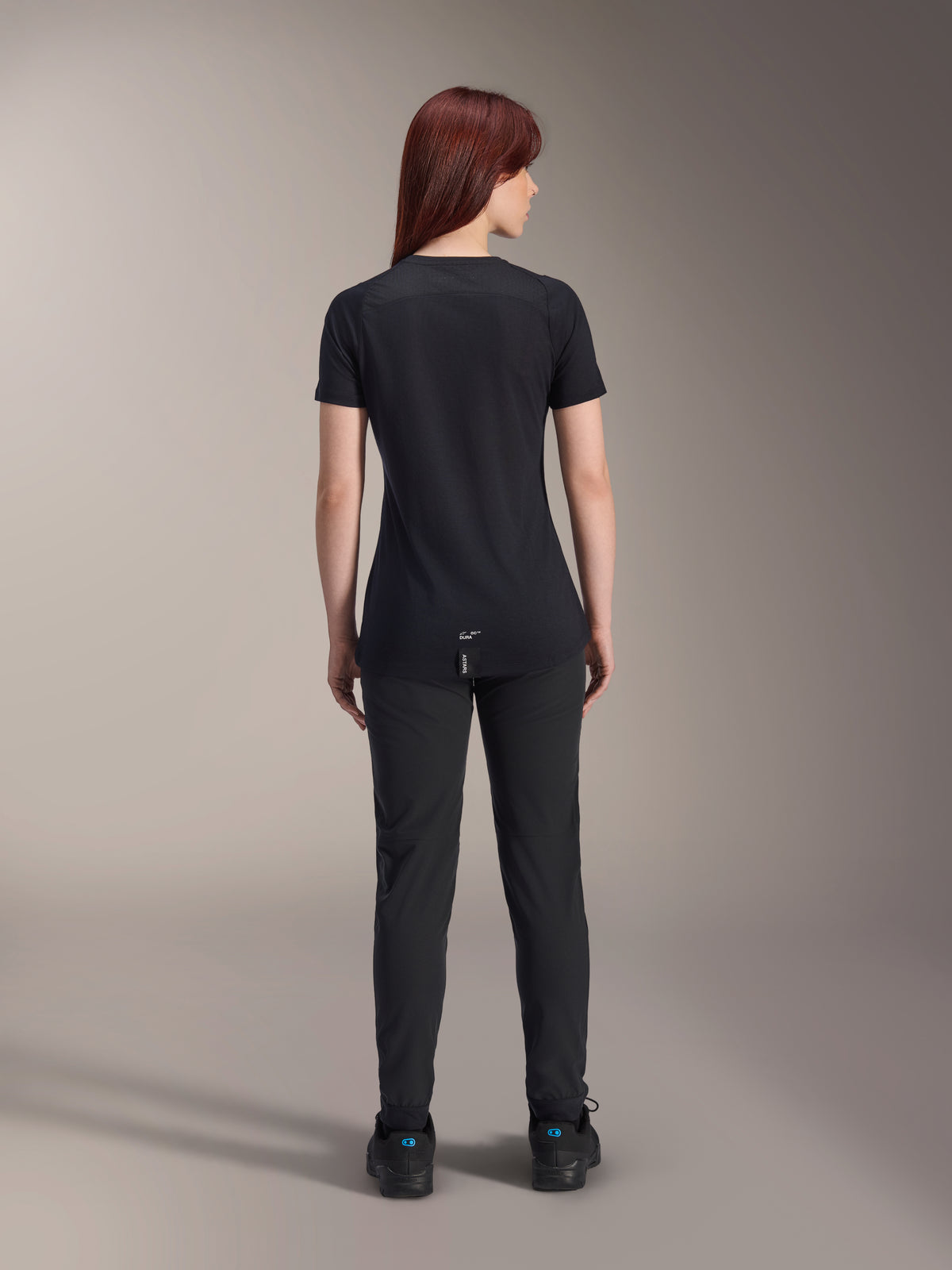 Alpinestars Damen Stella A-Dura Dri Switch Kurzarmtrikot, Schwarz, Rückansicht an einem Model, mit längerem abfallendem Saum und technischen Mesh-Einsätzen, kombiniert mit schwarzer Hose, Performance-MTB-Fahrradausrüstung