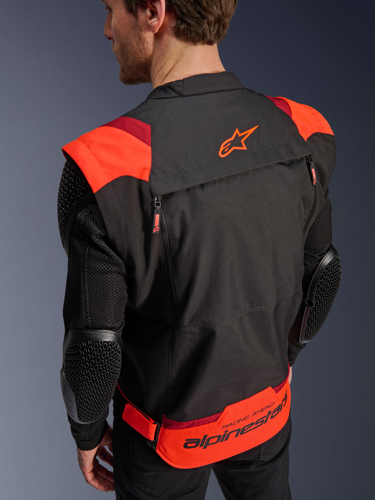 Alpinestars T-Stunt Air Jacke, Schwarz, Fluo-Rot und Dunkelrot, Rückansicht an einem Modell, zeigt Mesh-Ärmel mit wabenförmig strukturierten Ellbogenprotektoren, Alpinestars Stern-Logo auf dem oberen Rücken und markantes Branding am unteren Rücken mit 'RACING AHEAD'-Schriftzug, modulares Design mit sportlichem Schnitt für das Motorradfahren.