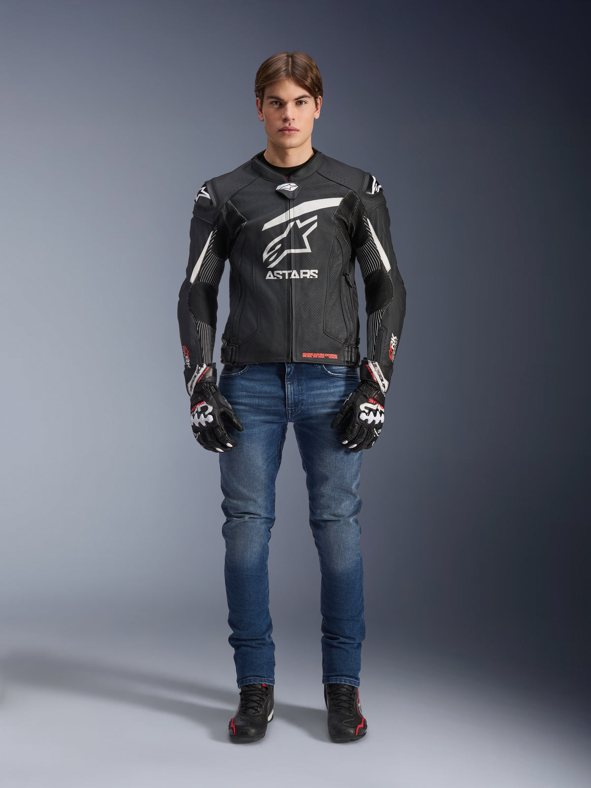 Männliches Model in Alpinestars GP Plus R V4 Rideknit Lederjacke in Schwarz und Weiß, kombiniert mit blauen Motorradjeans, schwarzen Motorradhandschuhen und schwarzen Motorradschuhen, großes weißes Astars-Logo auf der Brust, perforierte Lederbahnen und weiße Rennstreifen an den Ärmeln, Ganzkörperansicht im Stehen