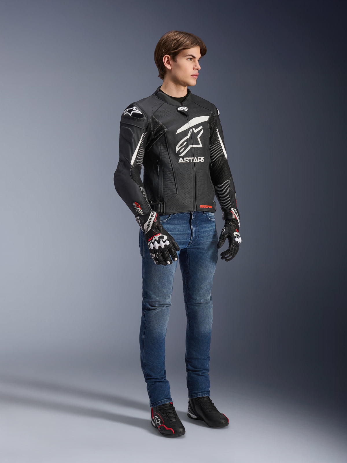 Alpinestars GP Plus R V4 Rideknit Lederjacke, Schwarz und Weiß, Motorrad-Lederjacke getragen von einem Modell mit blauen Jeans und schwarzen Handschuhen, mit einem markanten weißen Astars-Logo auf der Brust, perforierten Paneelen und weißen Akzenten an den Schultern.