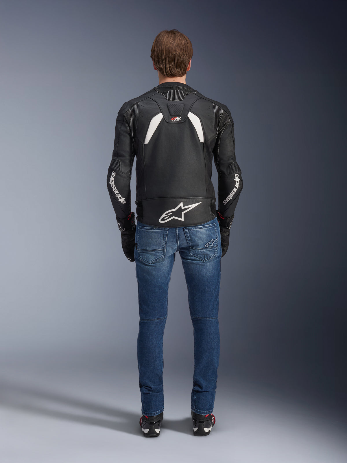 Rückansicht der Alpinestars GP Plus R V4 Rideknit Lederjacke, Schwarz und Weiß, mit einem großen weißen Logo im unteren Rückenbereich und Branding an den Ärmeln, getragen mit blauen Motorradjeans und schwarzen Stiefeln, zur Veranschaulichung der technischen Konstruktion und sportlichen Passform.