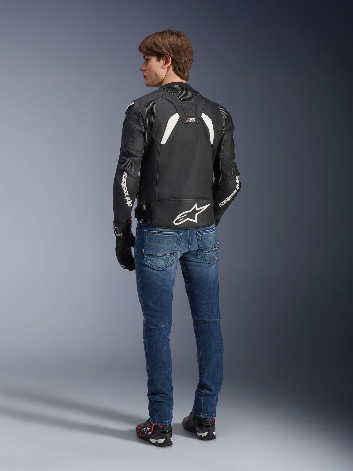 Alpinestars GP Plus R V4 Rideknit Lederjacke in Schwarz-Weiß, Rückansicht an einem Model, mit weißem Alpinestars-Branding an den Ärmeln und einem großen Stern-Logo auf dem unteren Rücken, kombiniert mit blauen Jeans und schwarzen Motorradschuhen, technische Motorradausrüstung.
