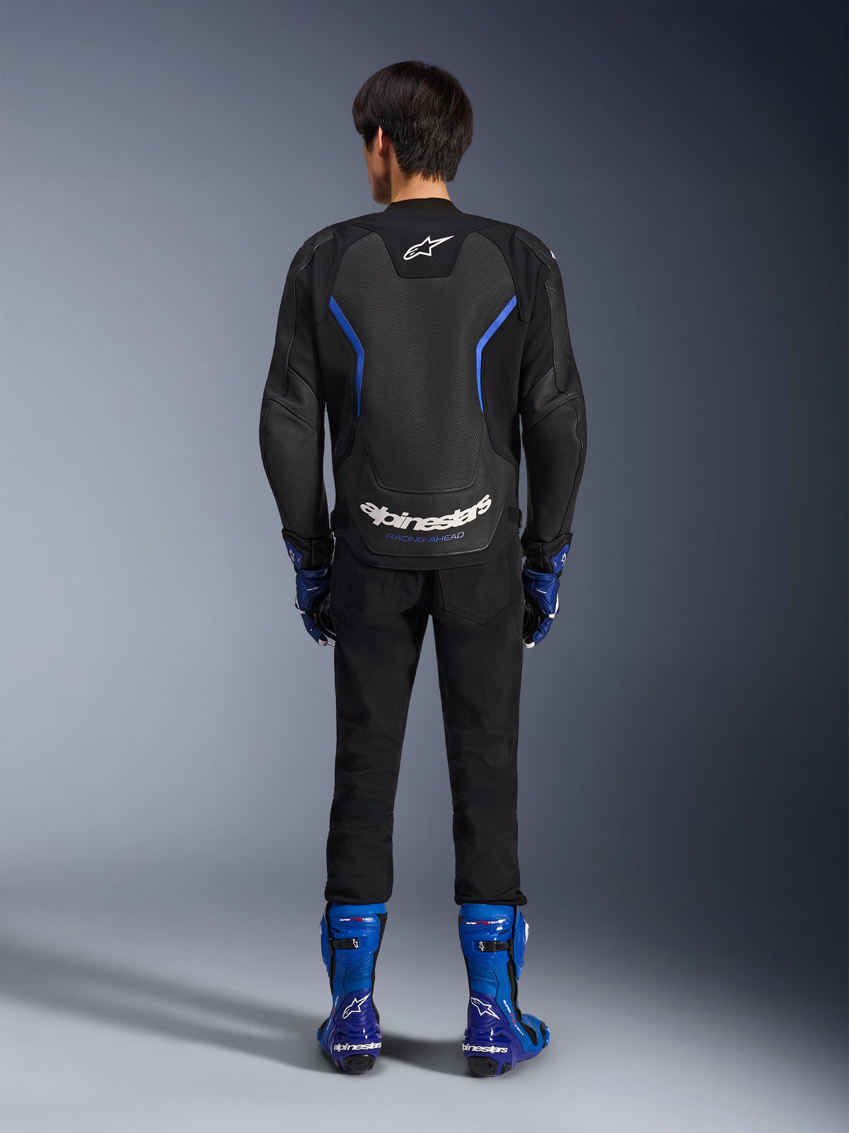 Alpinestars GP Force V2 Airflow Lederjacke, Schwarz Metallic-Blau, Rückansicht mit perforierten Lederbahnen und weißem Branding, getragen mit blauen Rennstiefeln und schwarzer Hose, Sport-Motorrad-Rennausrüstung