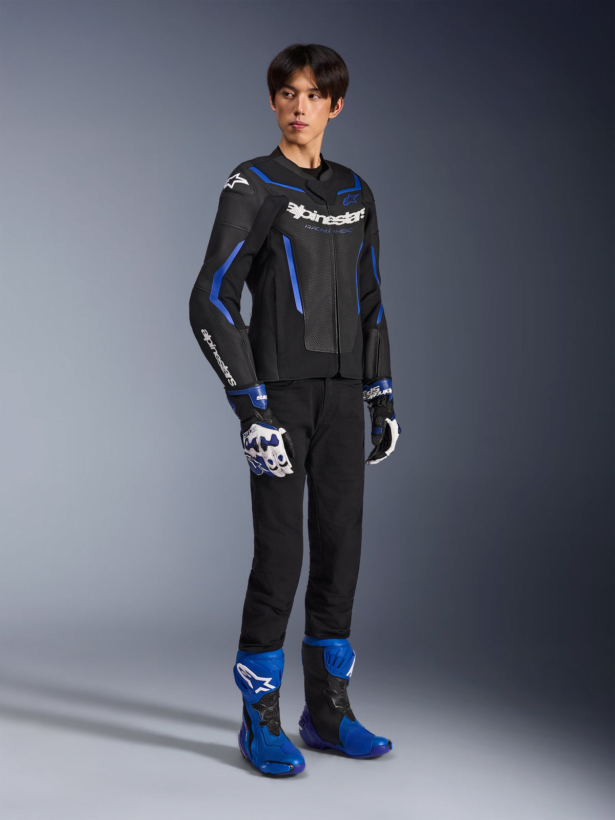 Männlicher Motorradfahrer in der GP Force V2 Airflow Lederjacke in Schwarz Metallic Blau, kombiniert mit passenden Rennhandschuhen und blauen Stiefeln, mit perforiertem Leder und markantem Alpinestars-Branding, professionelle Motorrad-Straßenausrüstung.
