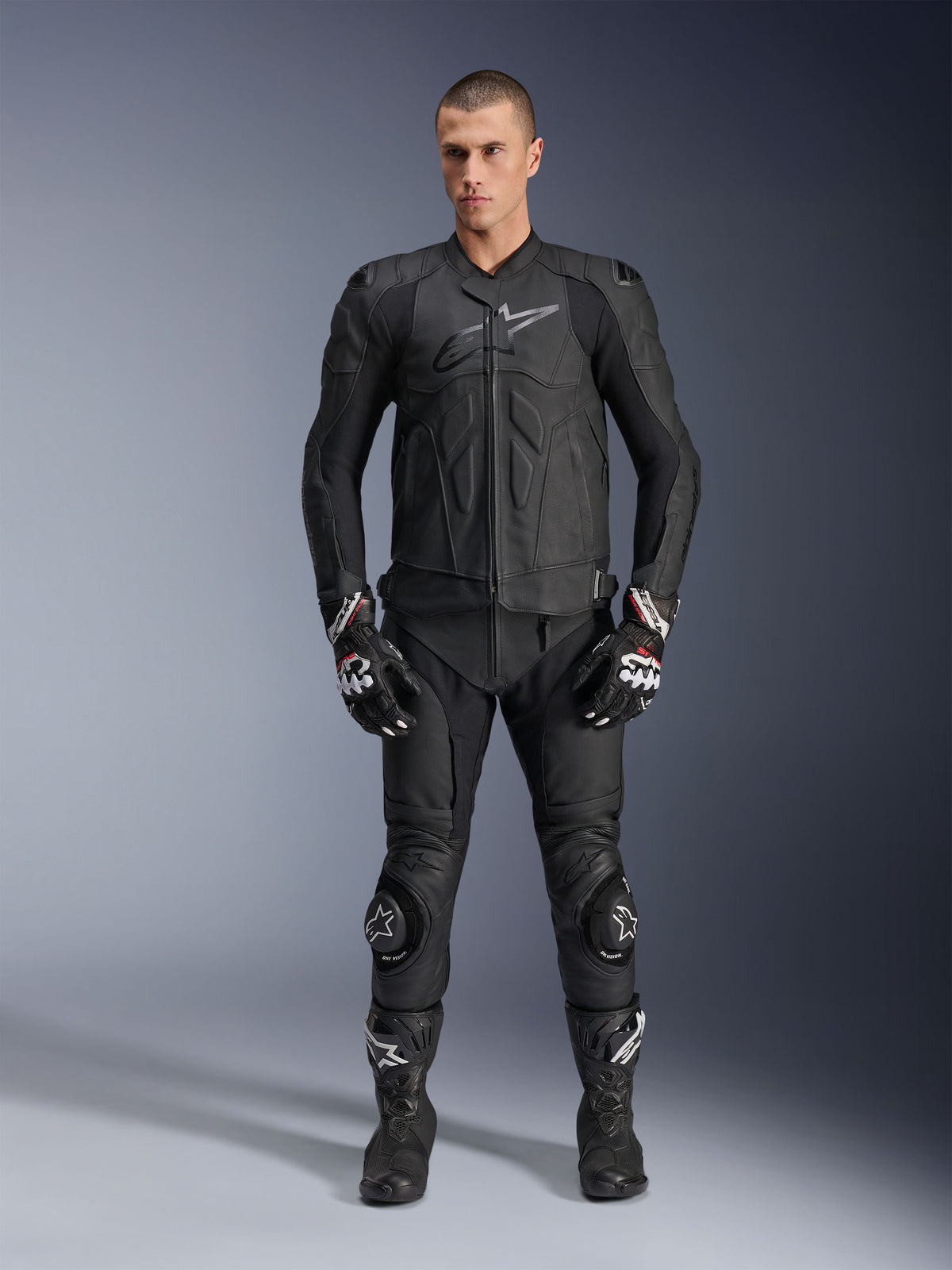 Alpinestars Dusk Lederjacke, Schwarz, getragen von einem männlichen Model als Teil eines kompletten, vom Rennsport inspirierten Outfits mit passender Lederhose, Handschuhen und Stiefeln, schlankes ergonomisches Design mit Protektoren an den Schultern und Markenlogo auf der Brust.
