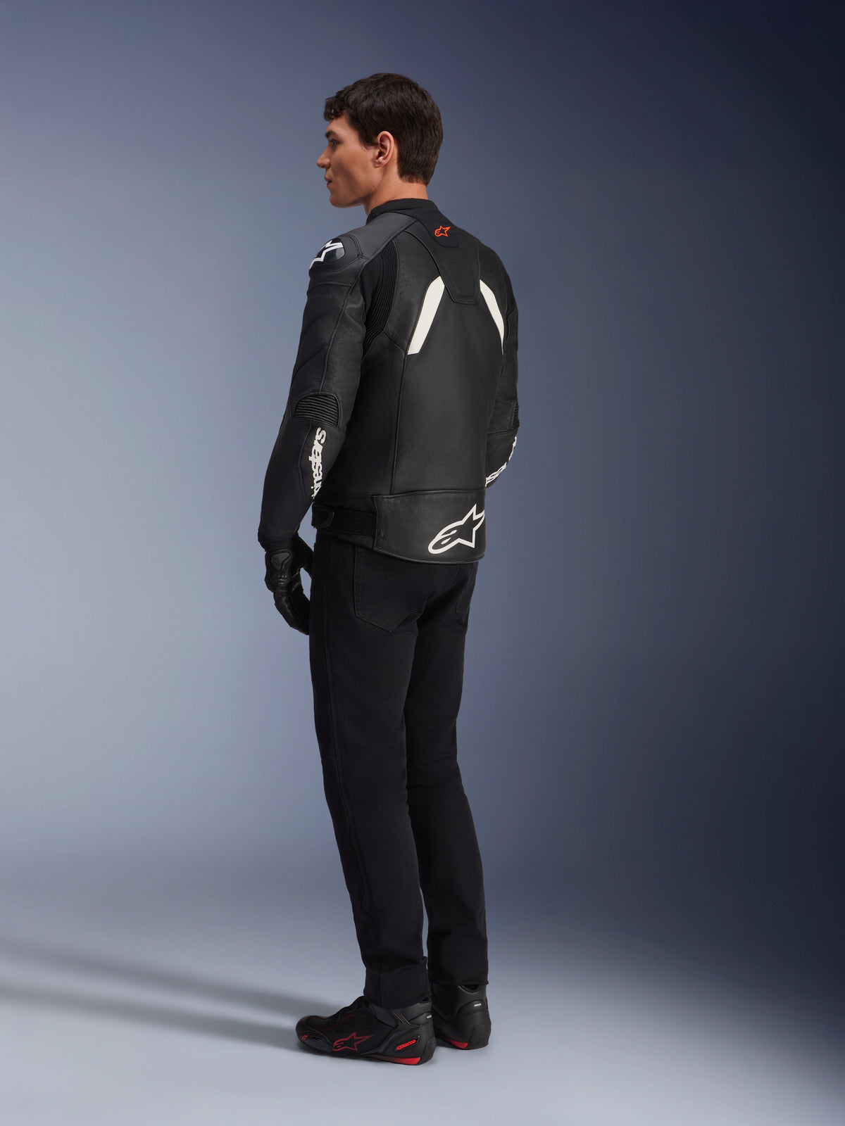 Alpinestars GP Plus V4 Leather Jacket, Sport Leather Jacket, Black and White, worn by a model in a rear-side profile view, featuring white Alpinestars logos on the lower back and sleeve, paired with black riding pants and technical boots for a sport performance look.