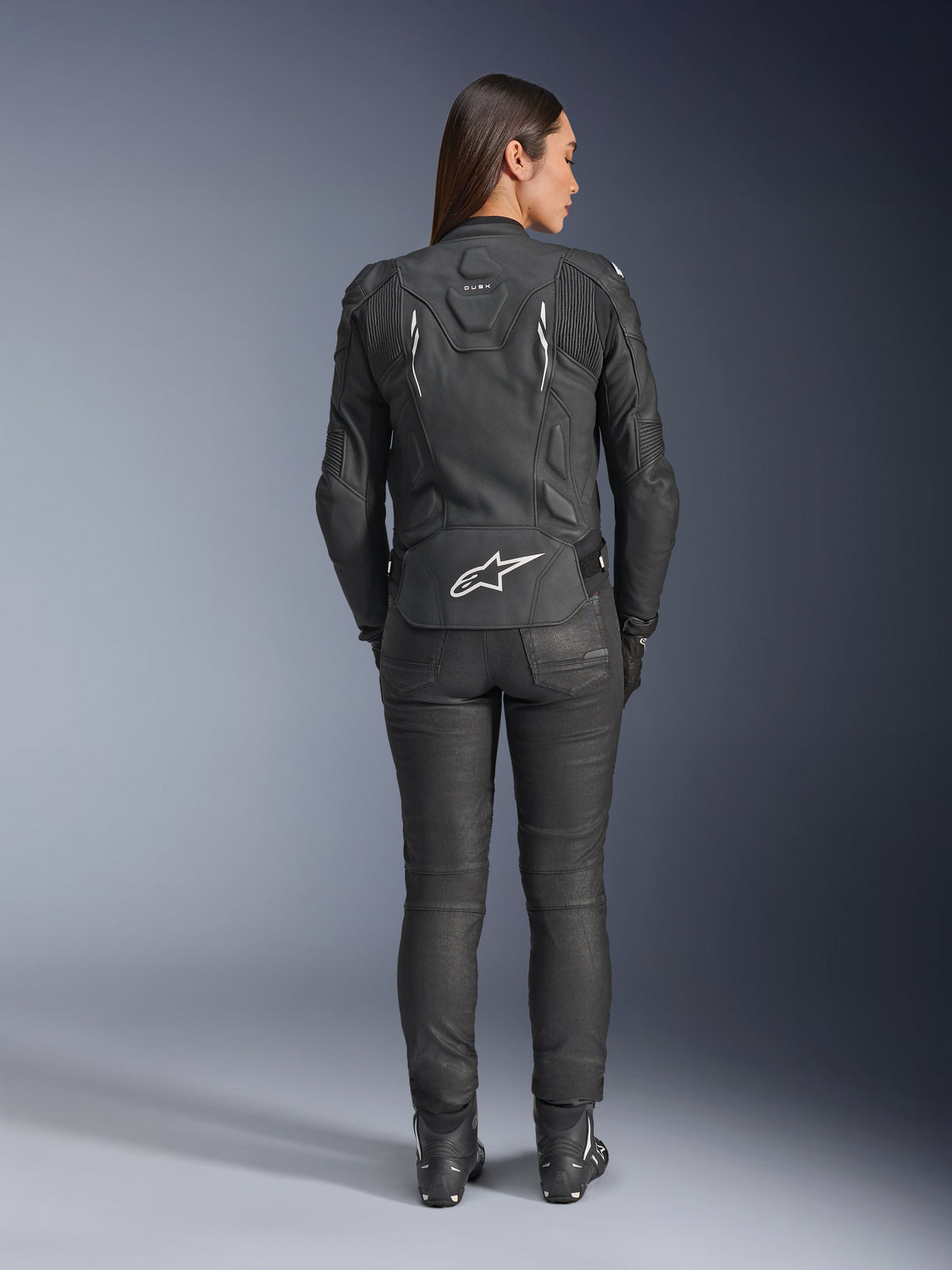 Alpinestars Woman Stella Dusk Leather Jacket, Black, Rear view featuring white Alpinestars logo on the lower back and 'DUSK' branding on the upper back, worn with black riding pants and boots, showcasing the contoured women's fit and protective leather construction.