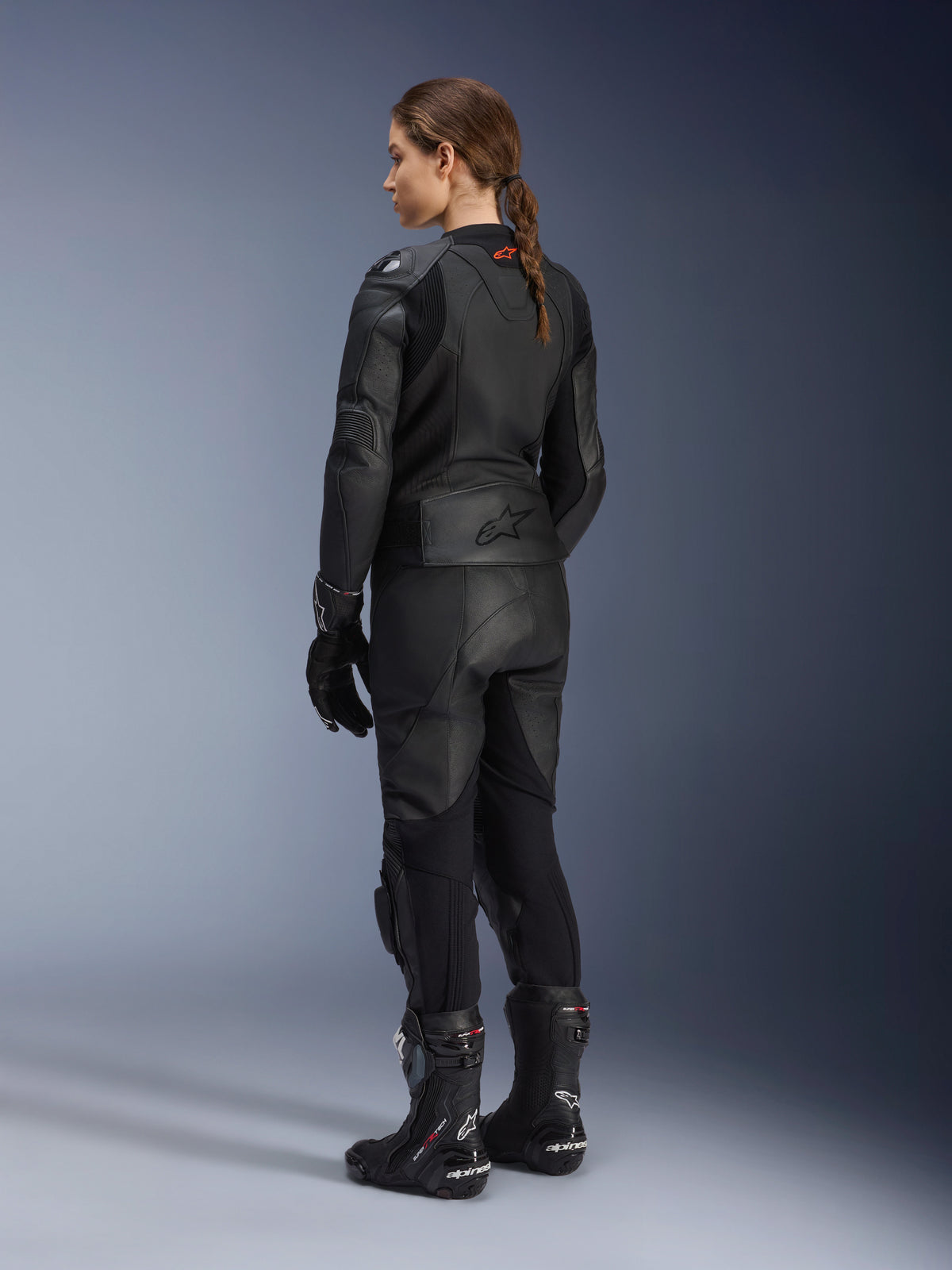 Women's Stella GP Plus V4 Leather Jacket, Black, full rear view, paired with black leather motorcycle pants, racing gloves, and boots, featuring embossed Alpinestars logo on the lower back and streamlined silhouette for sport riding