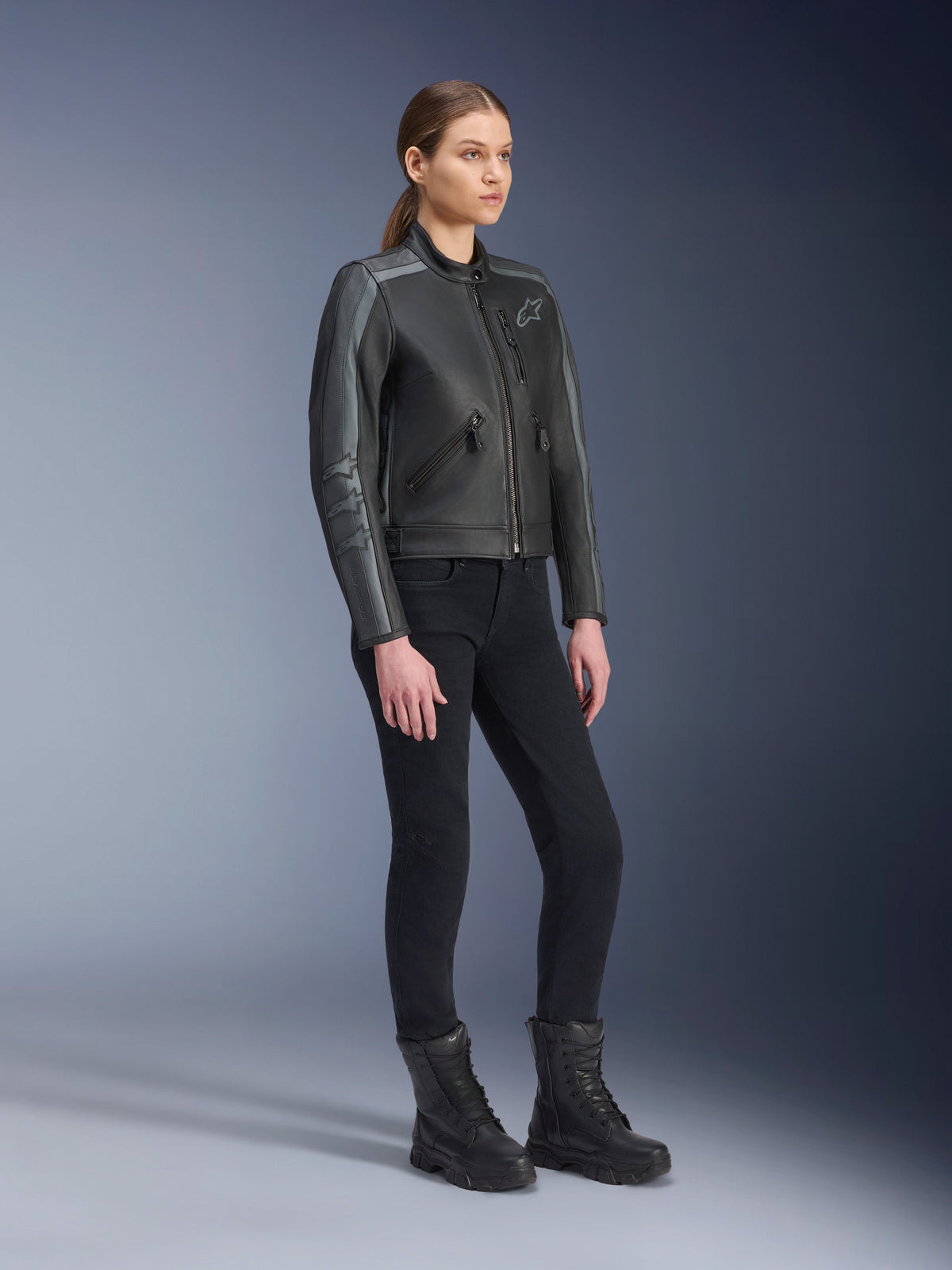 Alpinestars Stella Dyno Lederjacke, Damen-Motorradjacke, Schwarz, Tar Gray und Dunkelgrau, getragen mit schwarzer Motorradhose und Stiefeln, mit grauen Rennstreifen an den Ärmeln und Heritage-Lederdesign