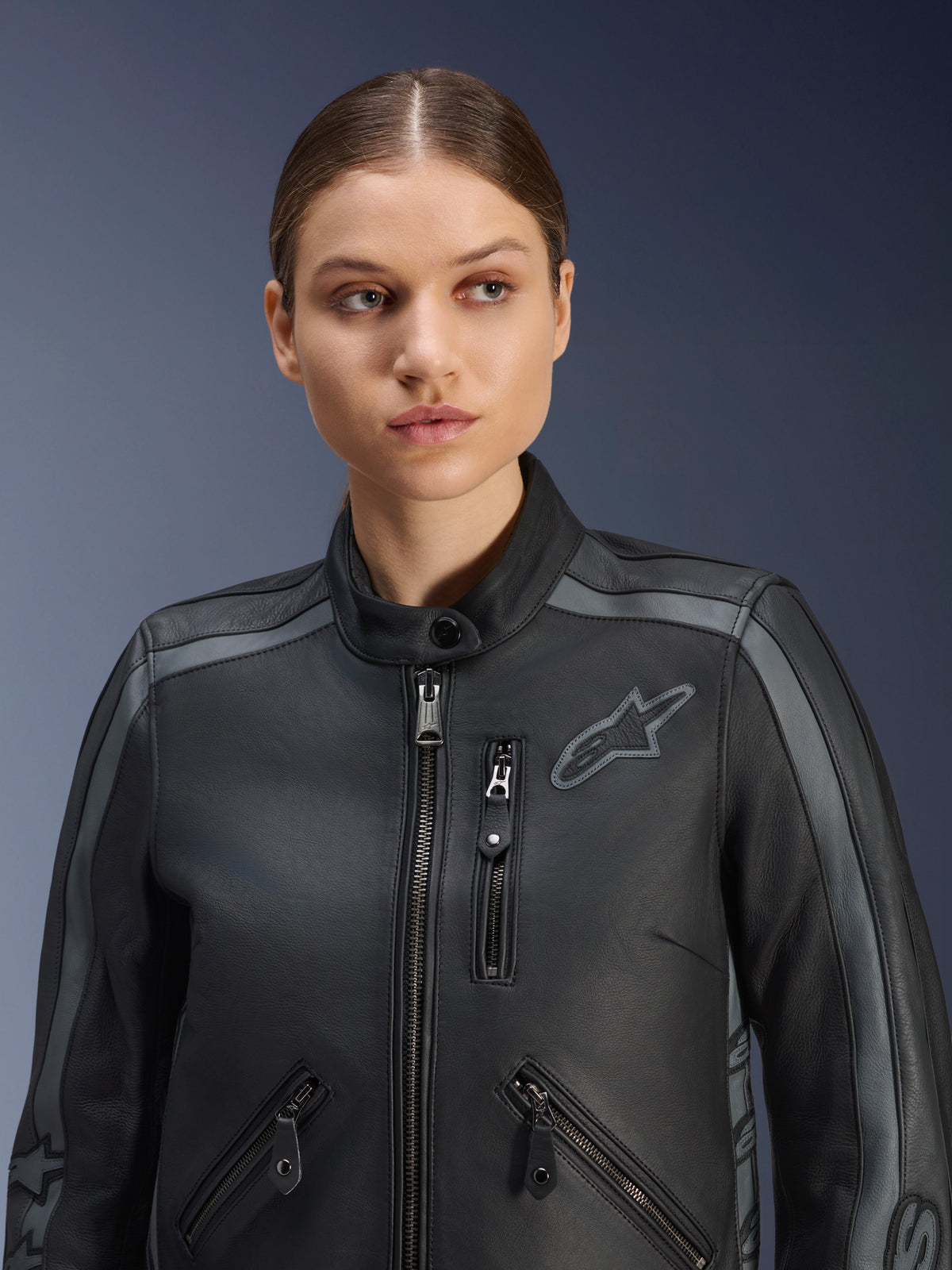 Weibliches Model trägt die Alpinestars Stella Dyno Lederjacke in Black Tar Gray Dark Gray, Premium-Rindsleder-Motorradjacke im Heritage-Design, mit grauen Schulterakzenten und mehreren Reißverschlusstaschen, speziell auf die weibliche Passform zugeschnitten.