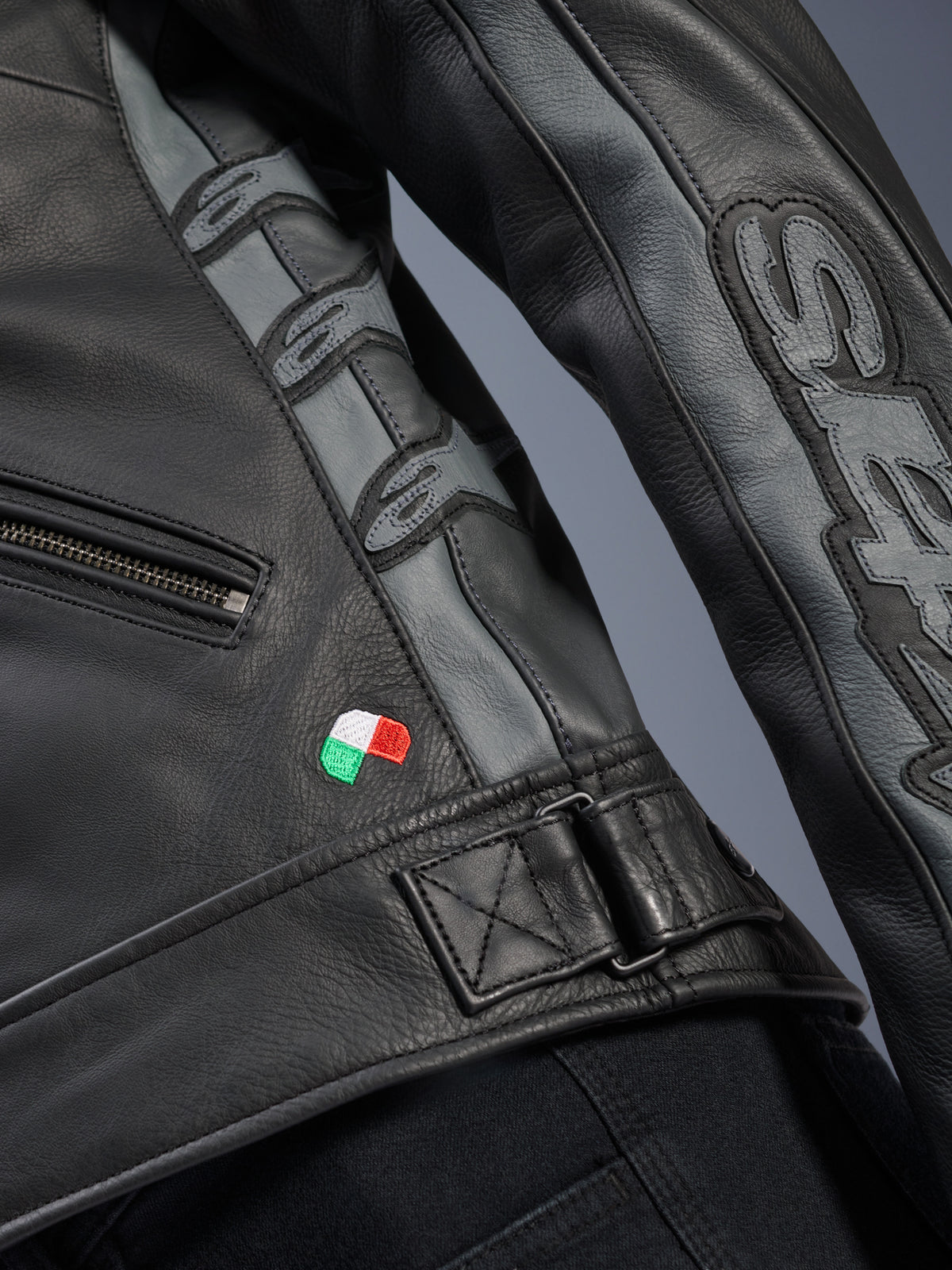 Alpinestars Stella Dyno Lederjacke, Detailansicht, schwarzes und teergraues Rindsleder, Nahaufnahme mit italienischer Flaggenstickerei, verstellbare Taillenschnalle, graue Ledereinsätze mit geprägten Alpinestars-Logos und Stella-Branding am Ärmel.