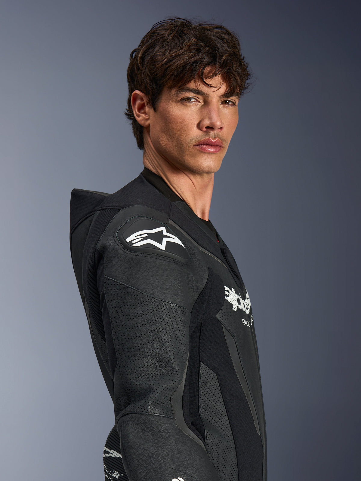 Männliches Model im Alpinestars GP Force V2 Lederkombi 1-teilig in Schwarz, Seitenprofilansicht mit Fokus auf das weiße Alpinestars-Sternlogo am DFS Lite Schulterprotektor, perforierte Lederpaneele und aerodynamischer Speed-Hump.