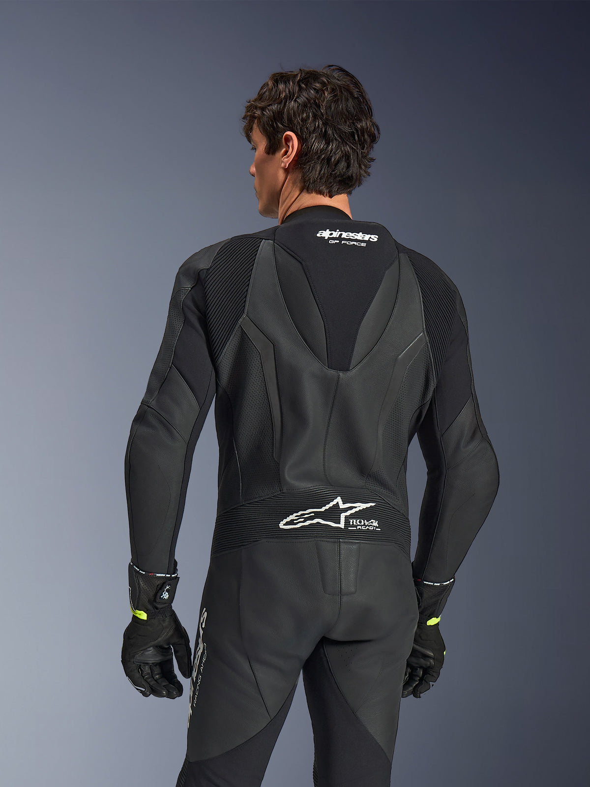 Alpinestars GP Force V2 Lederkombi, 1-teilig, Schwarz, Rückansicht zeigt perforiertes Leder und Akkordeon-Stretcheinsätze, weißes Alpinestars- und Tech-Air Ready-Branding am oberen und unteren Rücken, getragen mit schwarzen Handschuhen, Hochleistungs-Motorrad-Rennausrüstung