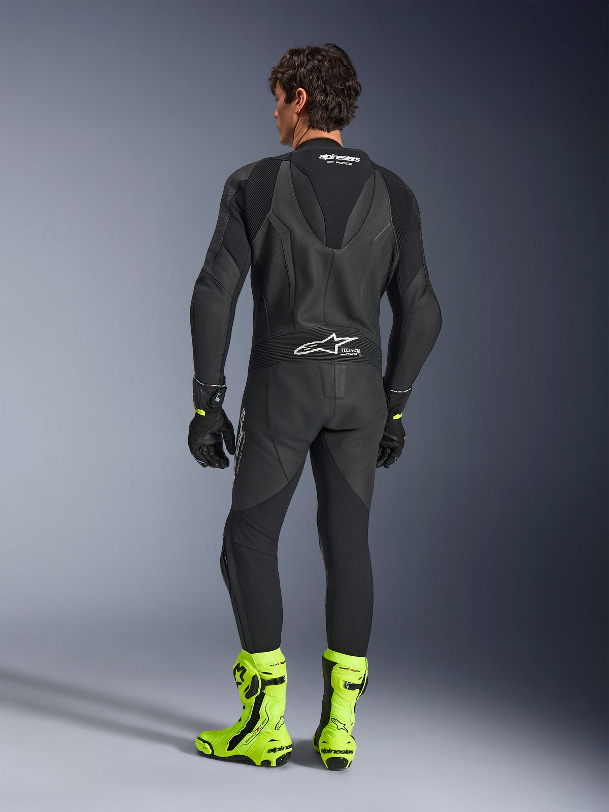 Alpinestars GP Force V2 Lederkombi - Einteiler, Schwarz, Rückansicht an einem Model, mit aerodynamischem Speed-Hump und Tech-Air Ready Branding, kombiniert mit schwarzen Handschuhen und leuchtend fluoreszierend gelben Rennstiefeln, professionelle Rennstreckenausrüstung