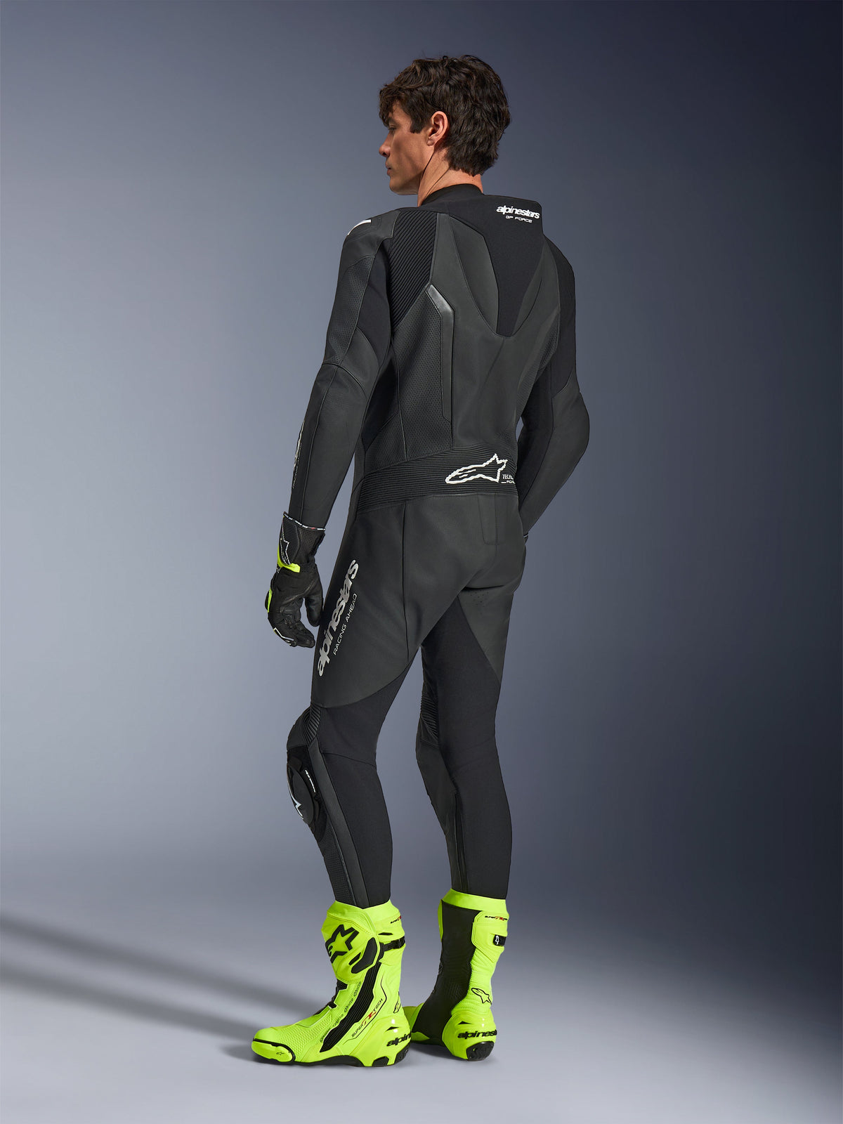 Alpinestars GP Force V2 Lederkombi, einteiliger schwarzer Motorrad-Rennanzug, Rückansicht mit aerodynamischem Speed-Hump und perforiertem Leder, kombiniert mit neongelben Rennstiefeln und schwarzen Handschuhen, professionelle rennstreckentaugliche Ausrüstung