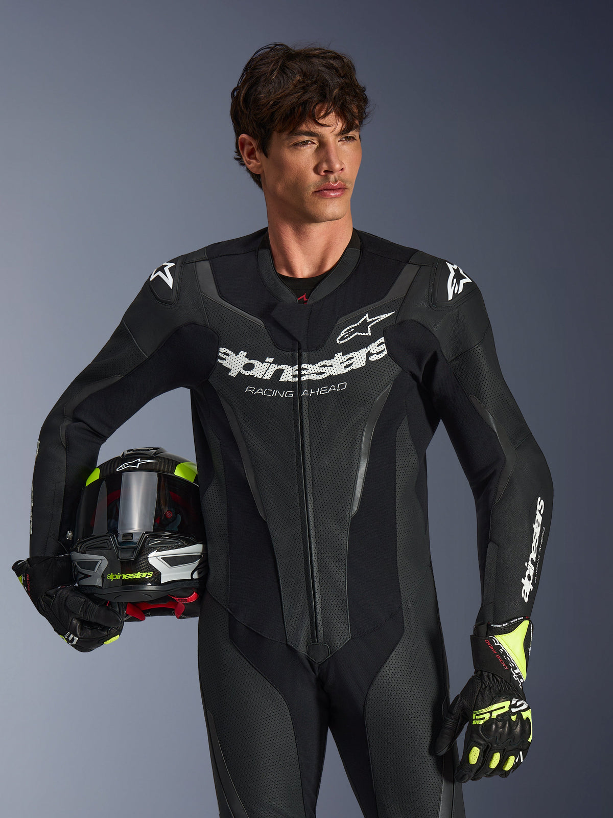 Alpinestars GP Force V2 Lederkombi, einteiliger schwarzer Motorrad-Rennanzug mit perforierten Ledereinsätzen und weißem Logo-Branding, getragen von einem Model mit Helm und passenden Handschuhen, Studiobeleuchtung