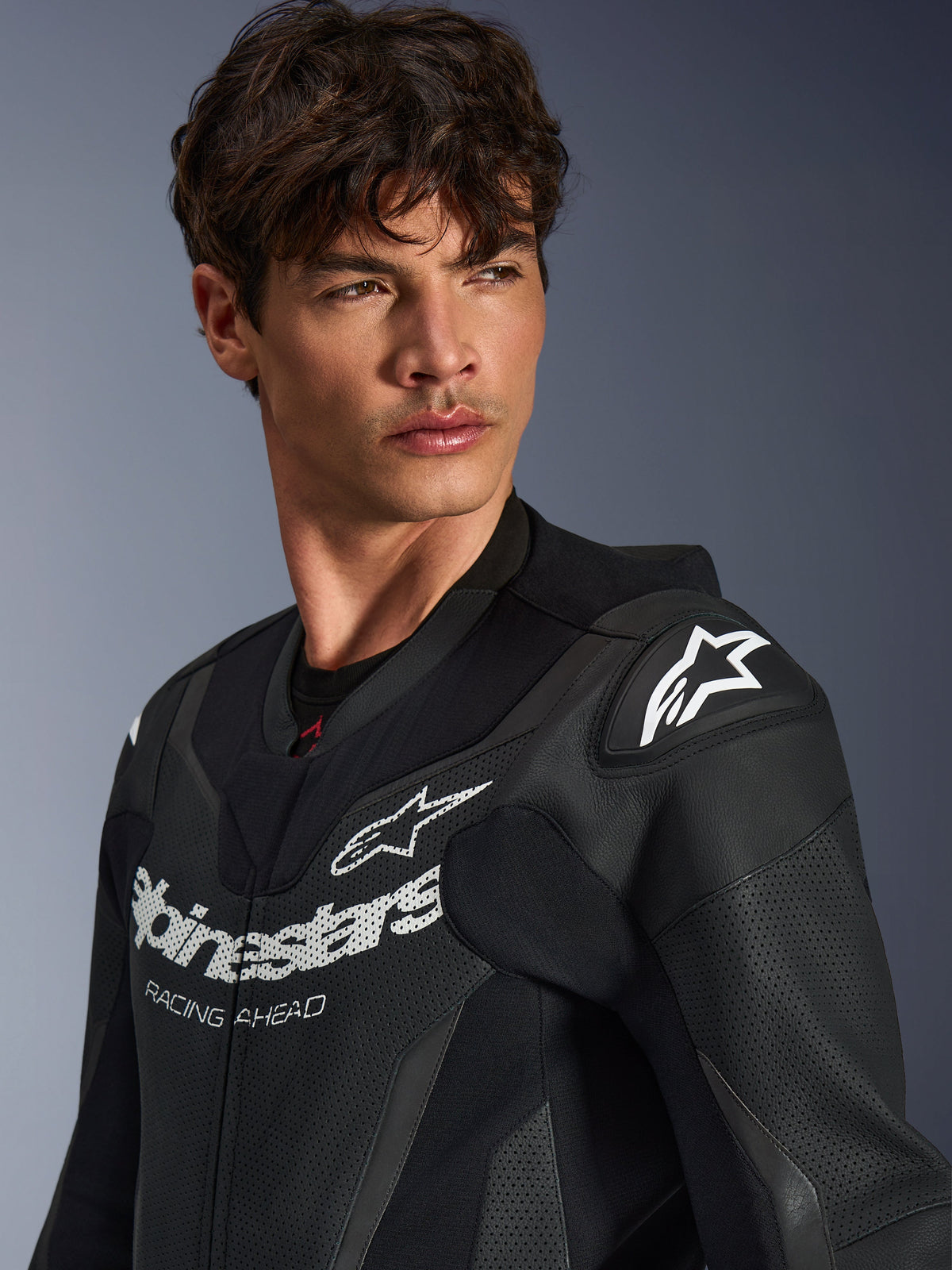 Nahaufnahme eines Mannes im schwarzen Alpinestars GP Force V2 Lederkombi, mit perforierter Lederkonstruktion, Alpinestars-Logo und 'RACING AHEAD'-Schriftzug auf der Brust sowie weißem Stern-Logo auf den Schulterschleifern.