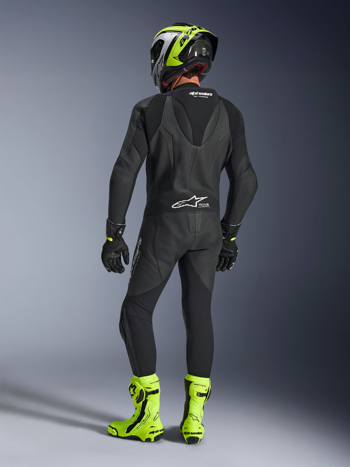 Rückansicht eines Fahrers im Alpinestars GP Force V2 1-teiligen Lederanzug in Schwarz, zeigt den aerodynamischen Speed-Hump und das Tech-Air Ready Branding, kombiniert mit einem schwarz-neongelben Helm und leuchtend gelben Rennstiefeln.