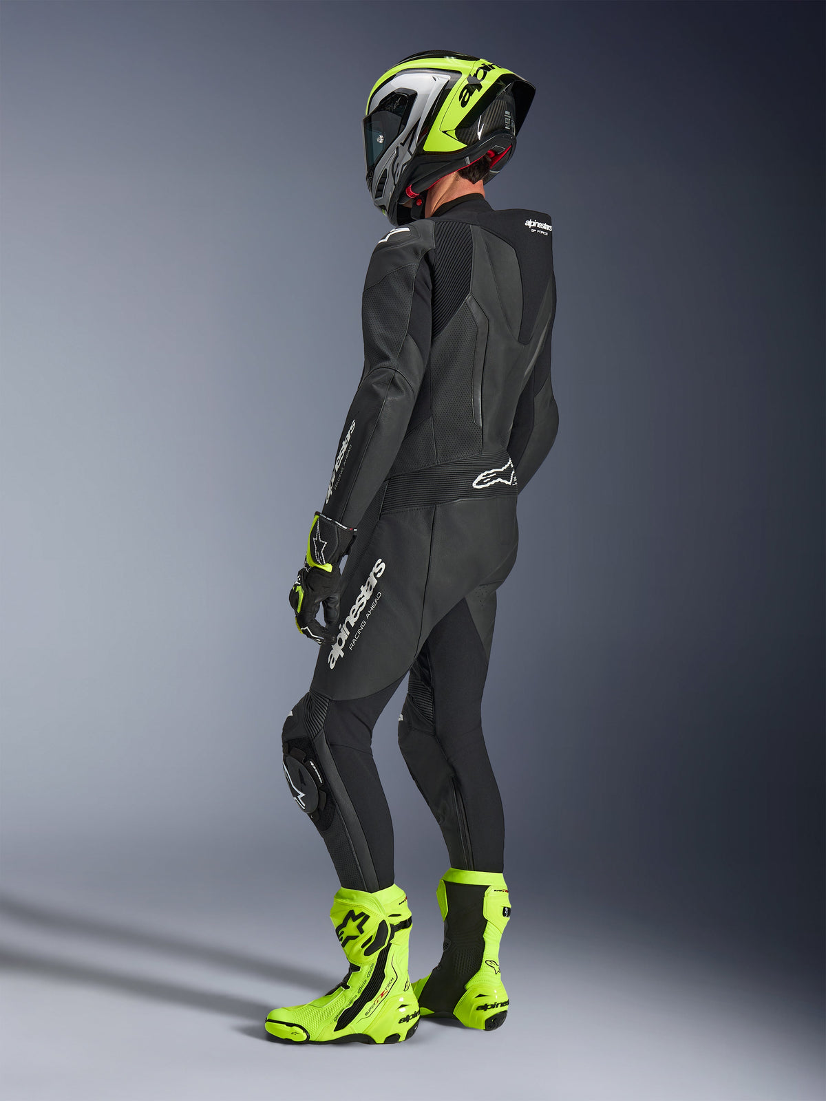 Motorradfahrer im Alpinestars GP Force V2 Lederkombi 1-teilig in Schwarz, Rückansicht mit weißem Branding am Oberschenkel und unteren Rücken, aerodynamischer Rückenhöcker, kombiniert mit einem neongelb-schwarzen Helm, passenden Rennhandschuhen und Stiefeln, professionelle Rennstrecken- und Sportfahrausrüstung.
