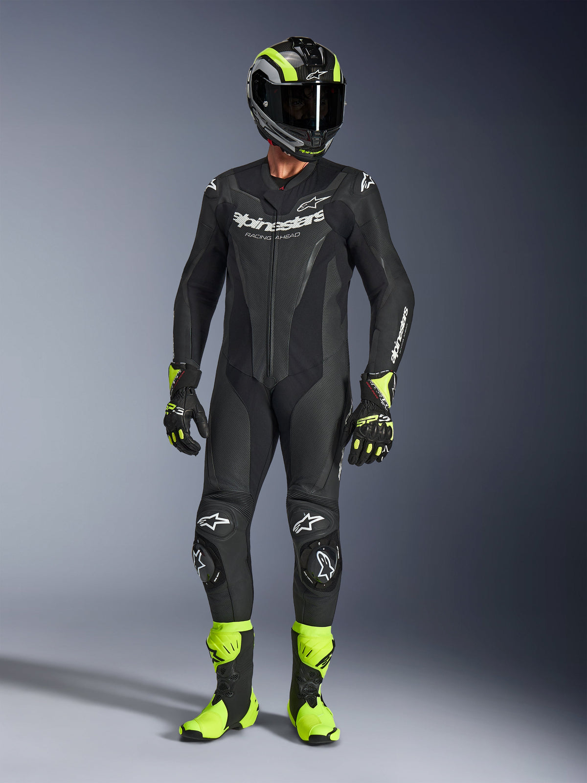 Motorradfahrer im Alpinestars GP Force V2 Lederanzug in Schwarz, einteiliger Lederanzug mit weißem Branding, kombiniert mit einem schwarz-fluorgelben Rennhelm, Handschuhen und Stiefeln, professionelle Rennausrüstung auf grauem Studiohintergrund