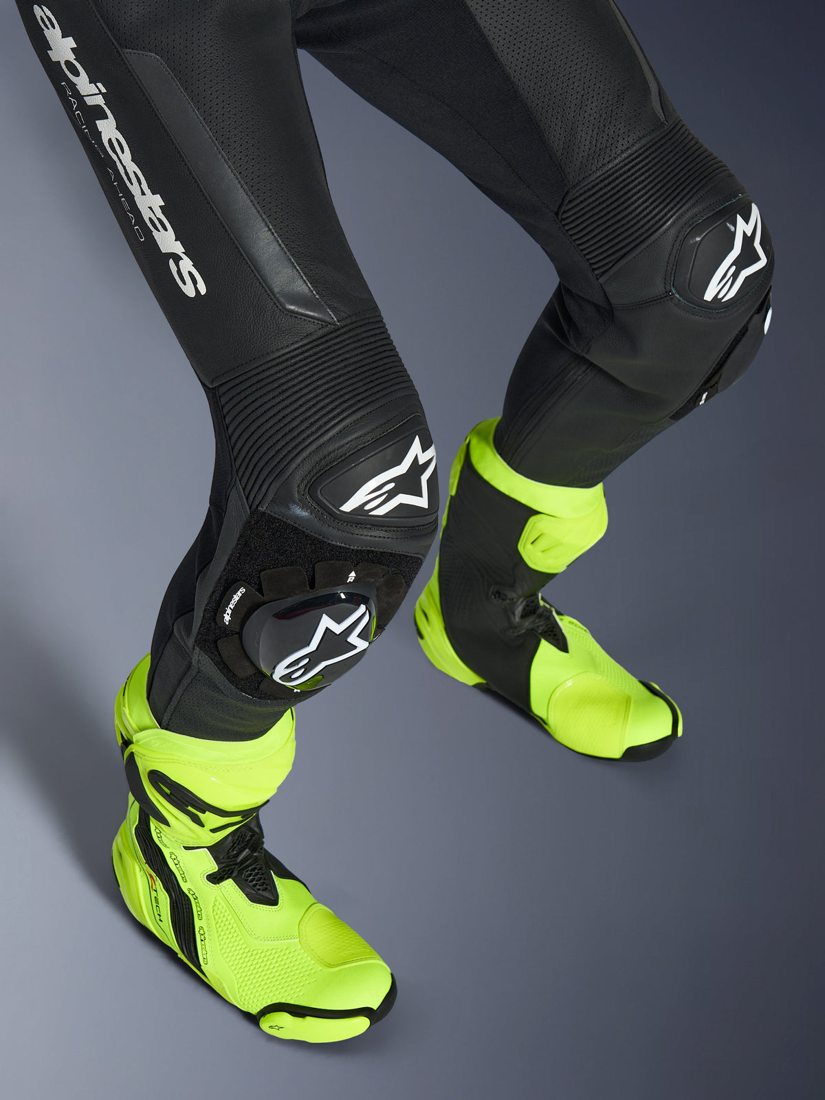 Alpinestars GP Force V2 Lederkombi, einteiliger schwarzer Lederanzug, Nahaufnahme der Beine und Knie mit weißen Logo-Schleifern, Akkordeon-Stretcheinsätze, getragen mit leuchtend neongelben Rennstiefeln, Hochleistungs-Motorradrennausrüstung